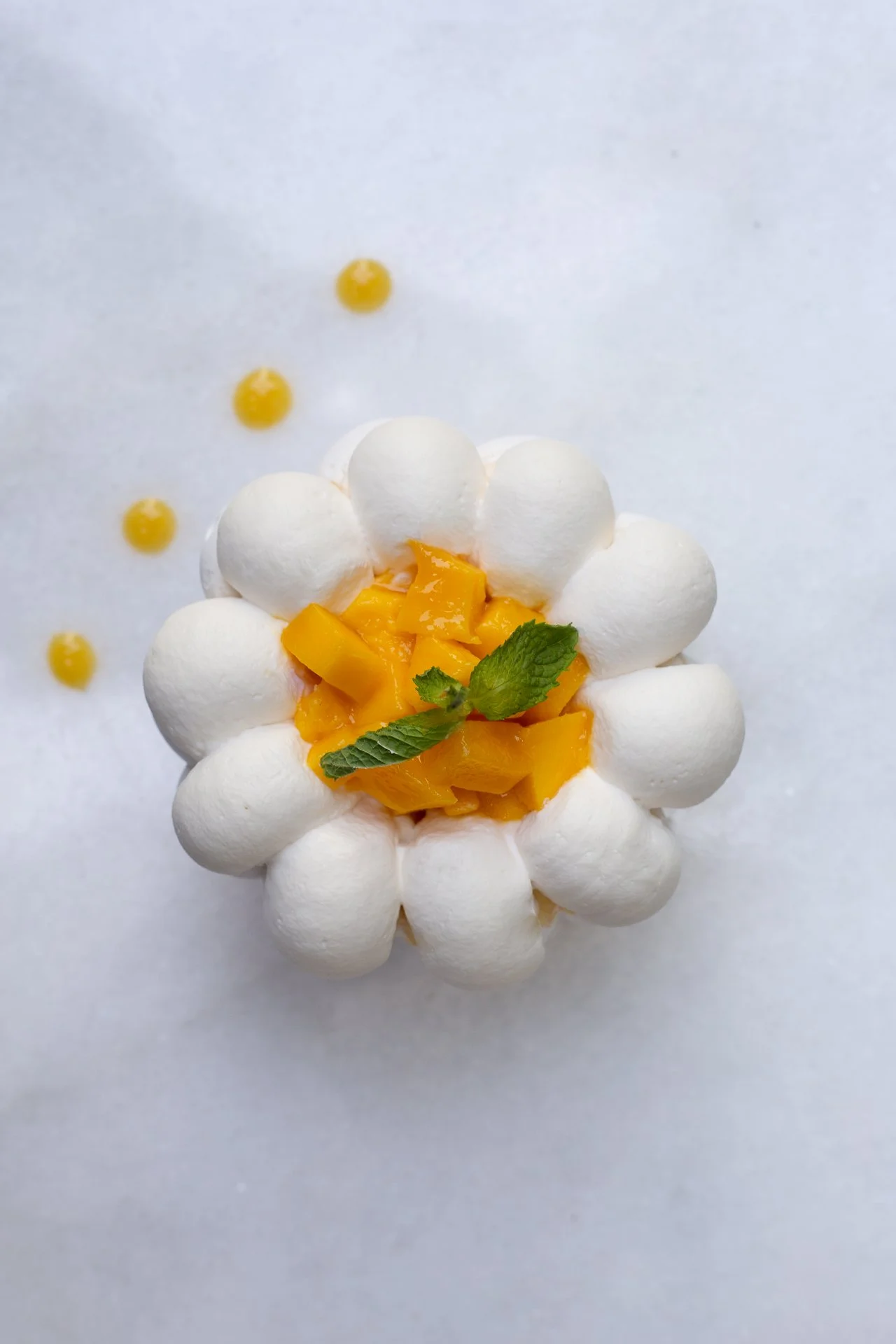 A dessert with white meringue-like dollops arranged in a circle, topped with chopped mango pieces and a small green mint leaf, on a white surface. Pavlova Mangue et Passion (Mango & Passion Fruit Pavlova).