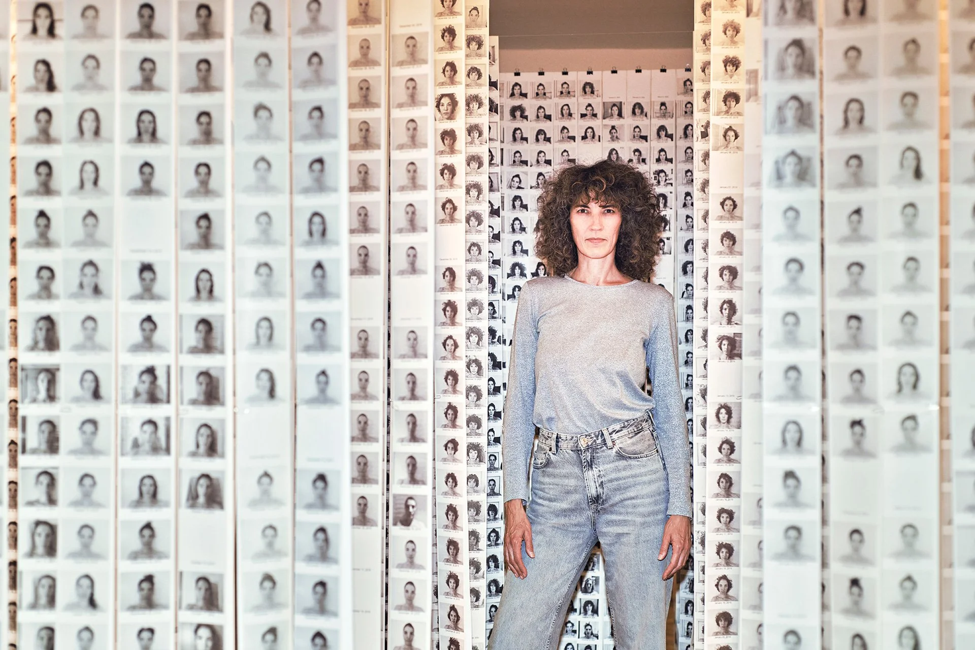 A curly-haired woman in a gray top and jeans stands among walls of tiny portrait photos, creating a striking, contemplative fashion portrait.