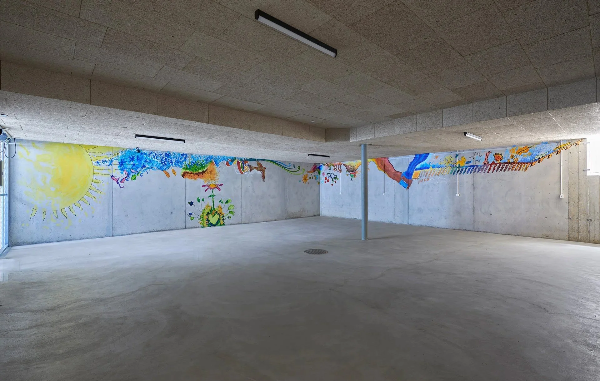 An empty, low-ceilinged concrete room has a bright, childlike mural of a sun, plants, and abstract shapes wrapping along the walls; quiet and spacious.
