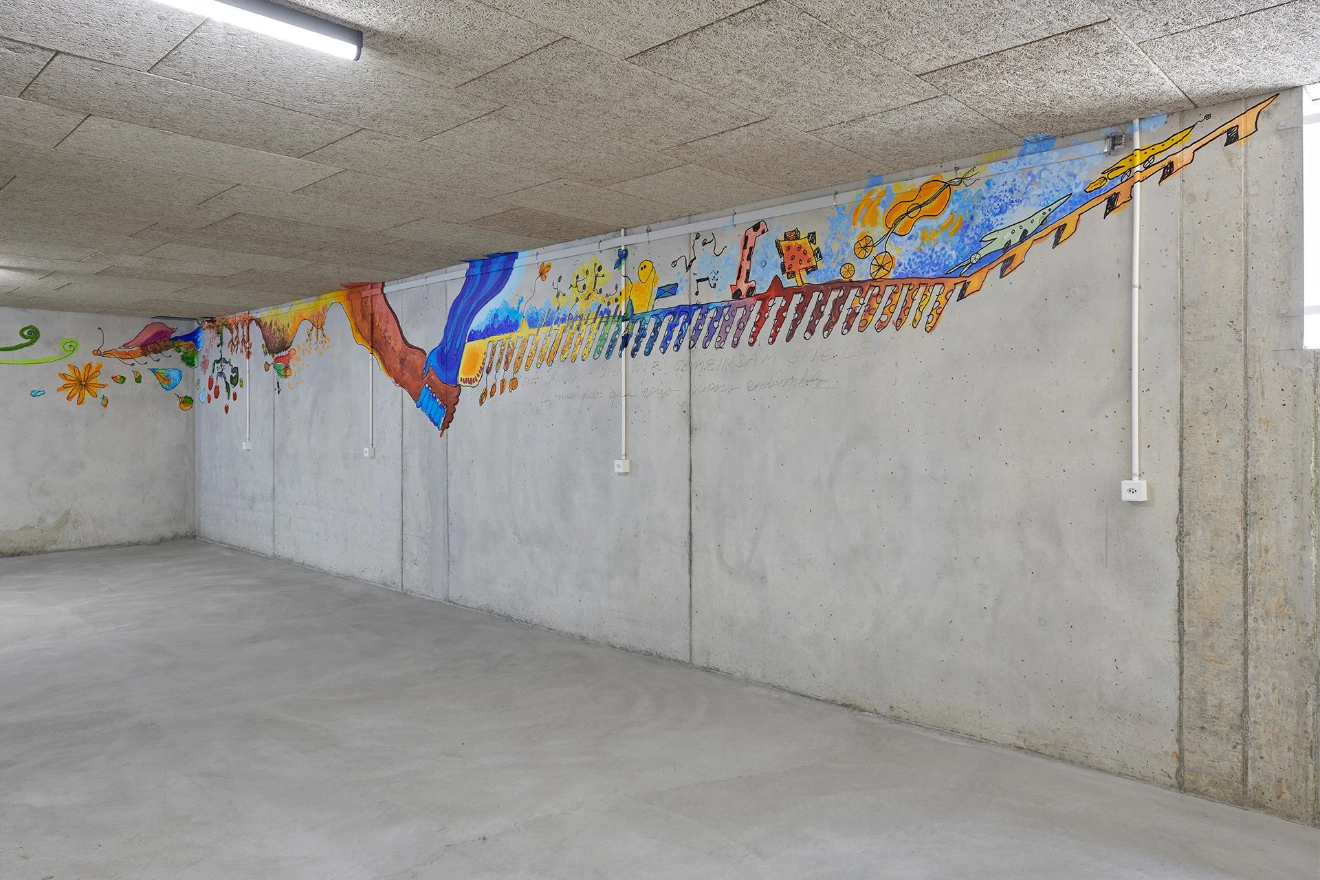 A colorful mural of abstract shapes and figures stretches across a bare concrete basement wall beneath fluorescent lighting, creating a playful contrast with the stark, quiet space.