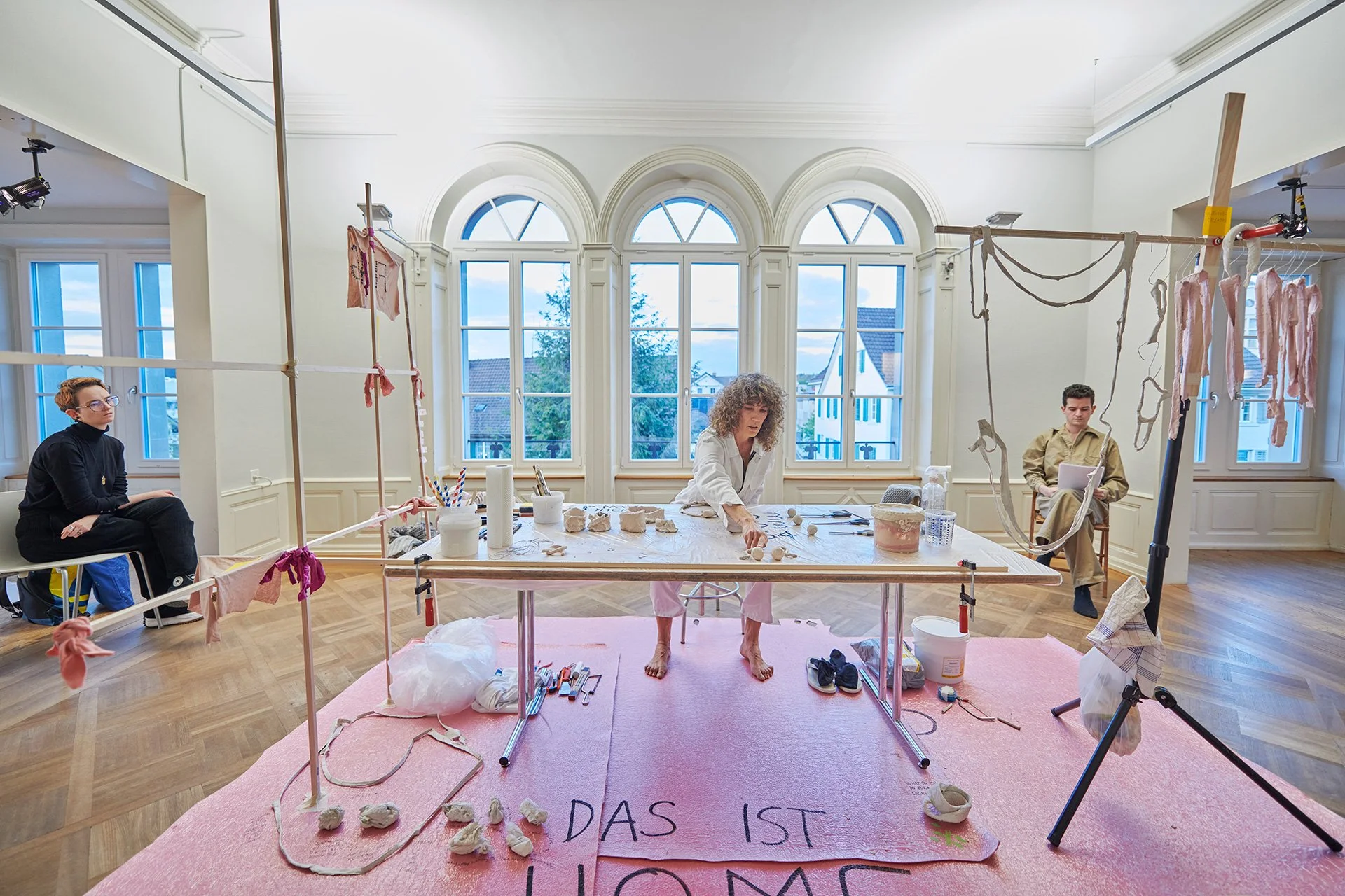 An airy art studio with three people working around a long table, tools and hanging materials, and a pink floor installation beneath arched windows.
