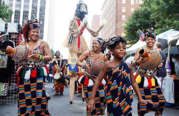 Dancers in kente cloth with shekeres and stilt walker parade down Fayetteville Street