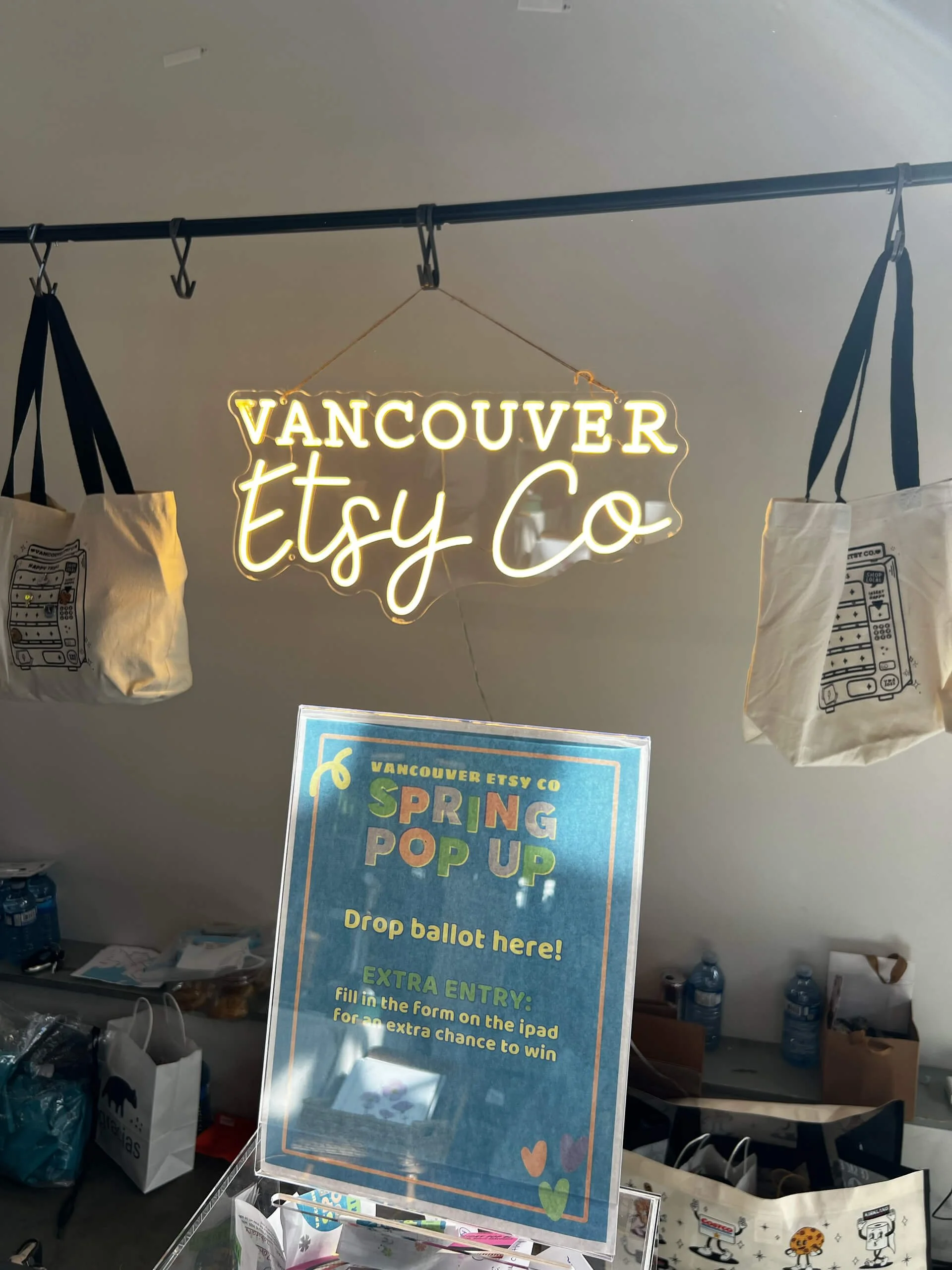 Neon sign that reads "Vancouver Etsy Co" hanging from the ceiling. Various tote bags are displayed on a rack, and a sign for an event called "Spring Pop Up" is visible below the neon sign. There are water bottles and other items on a table against th