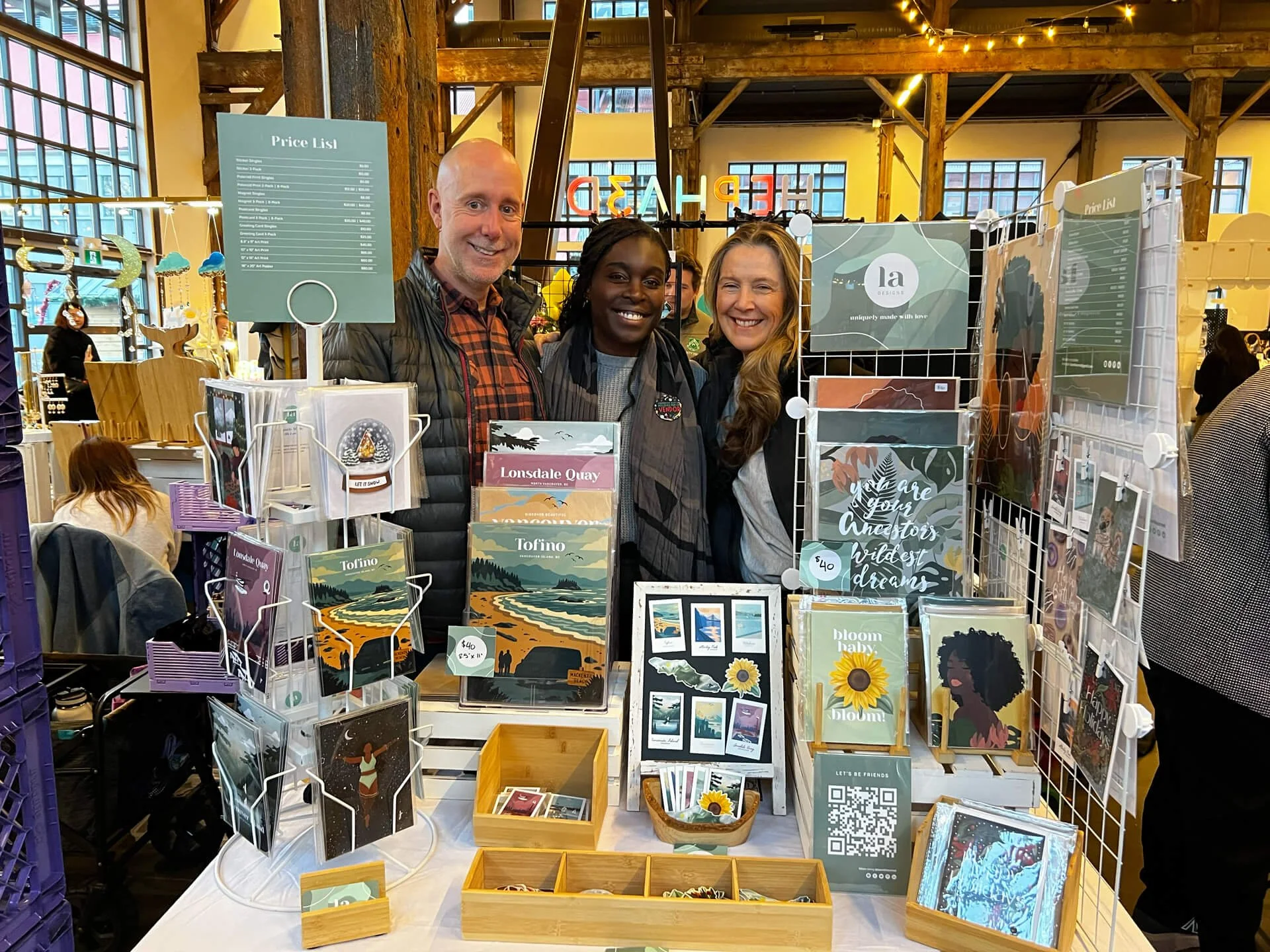 Three smiling people standing behind a table at a craft fair or market booth with art prints and greeting cards for sale, inside a well-lit industrial-style building with large windows and exposed wood beams.