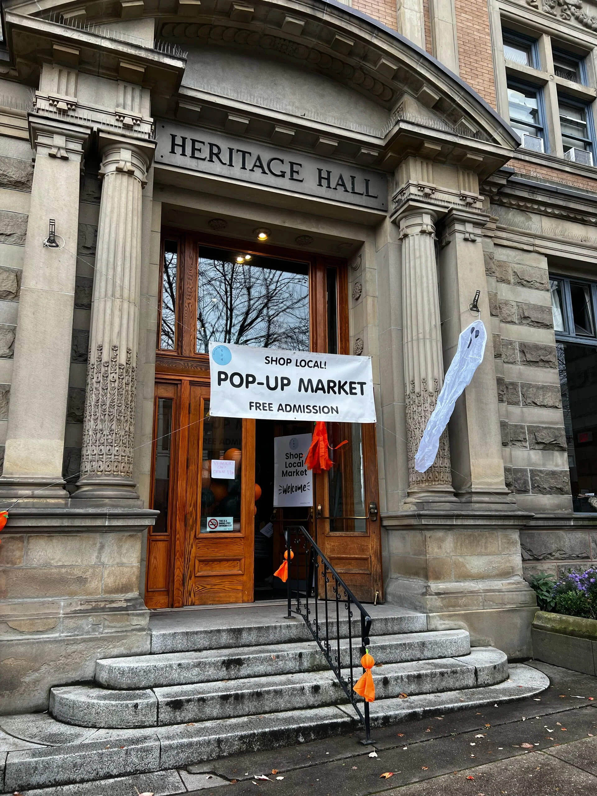 The entrance of Heritage Hall, a historic building with stone steps and ornate architectural details. A banner announces a pop-up market with free admission, and Halloween decorations, including a hanging ghost and orange pumpkins, are visible.