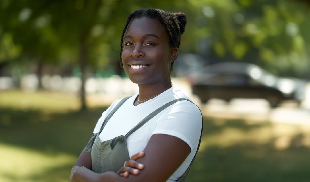 The female designer behind la designs with dark hair styled in twists, smiling, standing outdoors with arms crossed, wearing a white shirt and overalls.