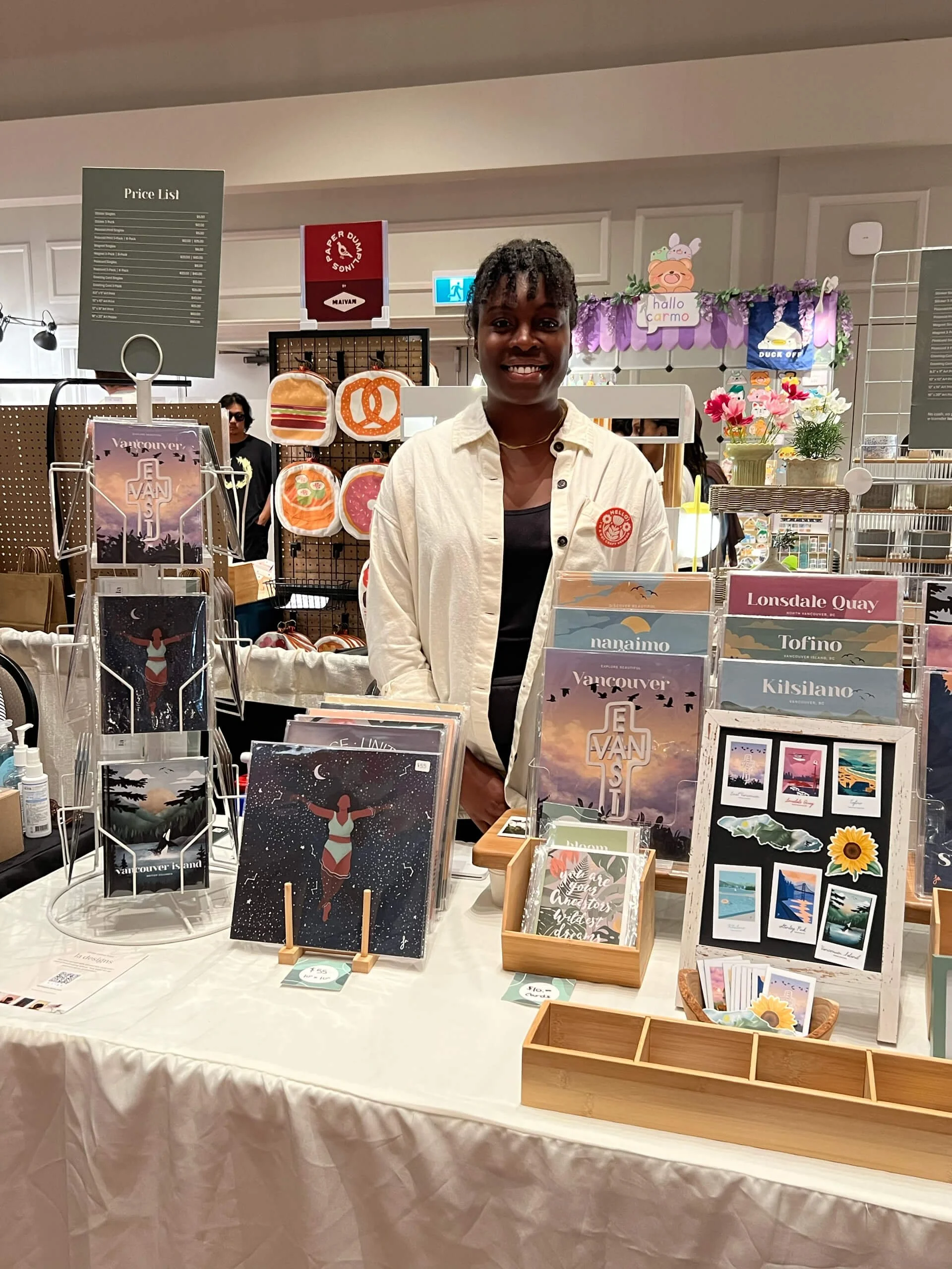 A smiling young woman standing behind a display table at a craft fair or market, showcasing various colorful postcards and printed artworks with nature and travel themes. The background features shelves with more items, signs, and decorative elements