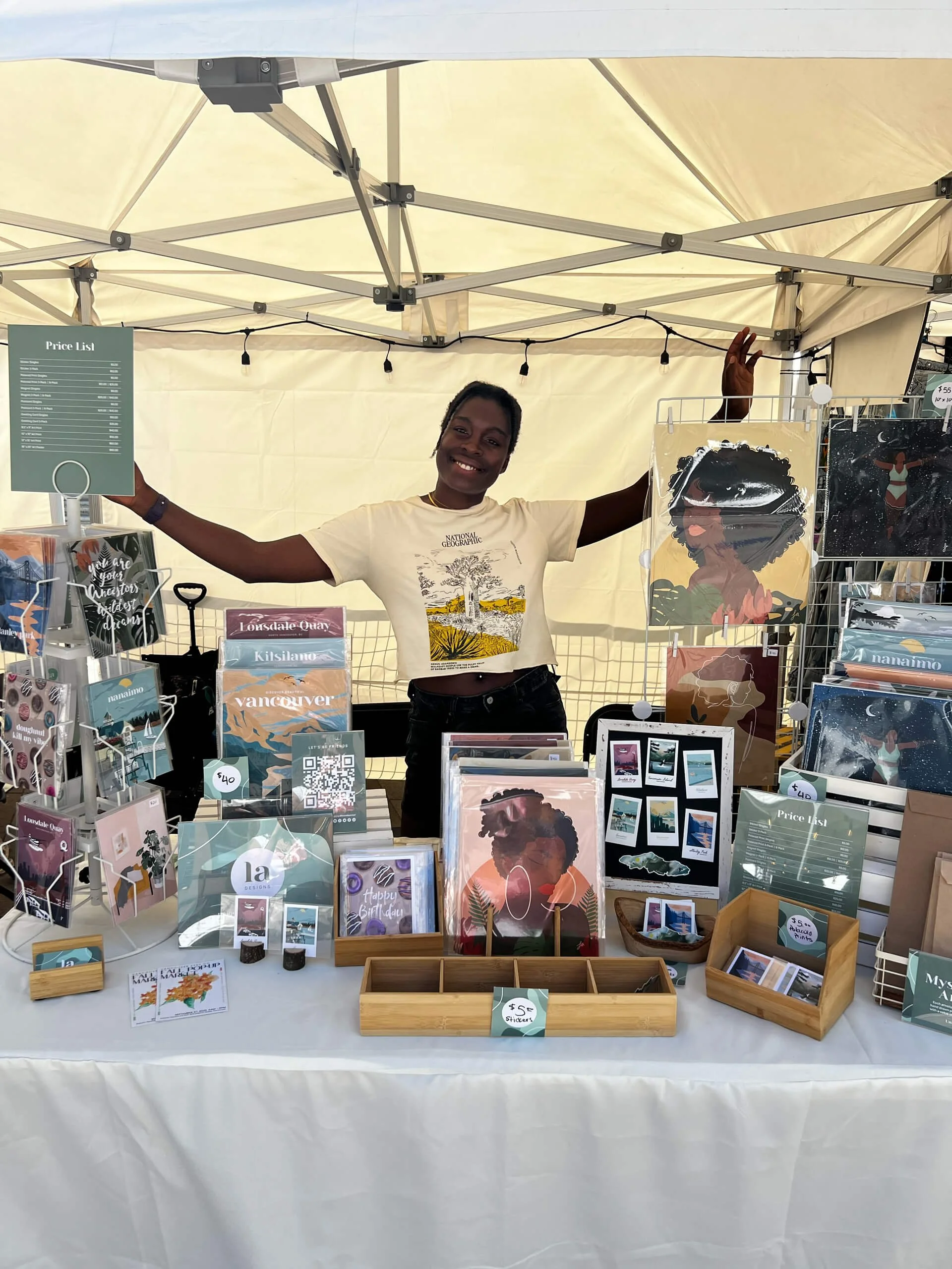 A woman standing behind a display table at an art or craft fair, smiling and waving. The table has various illustrated prints, greeting cards, and small art pieces, with some price tags visible. The tent overhead is supported by metal poles, with str