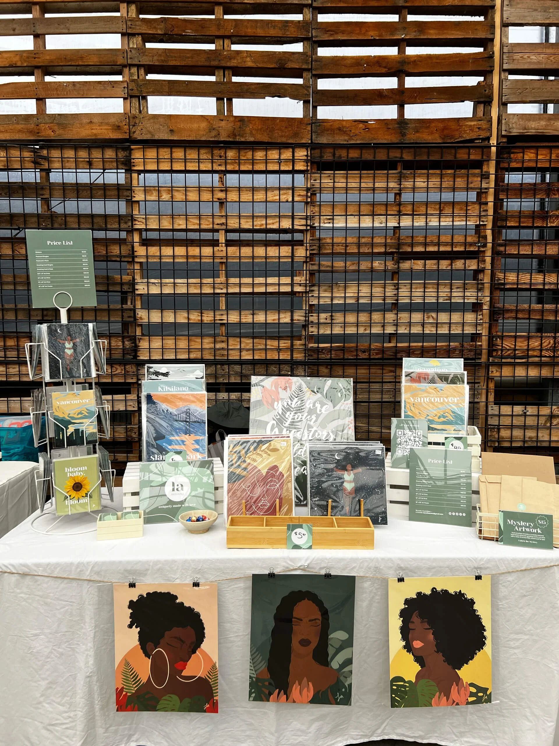 A display table at an art fair features colorful posters of Black women with natural hairstyles and jewelry, alongside small items and a greens-themed art print.