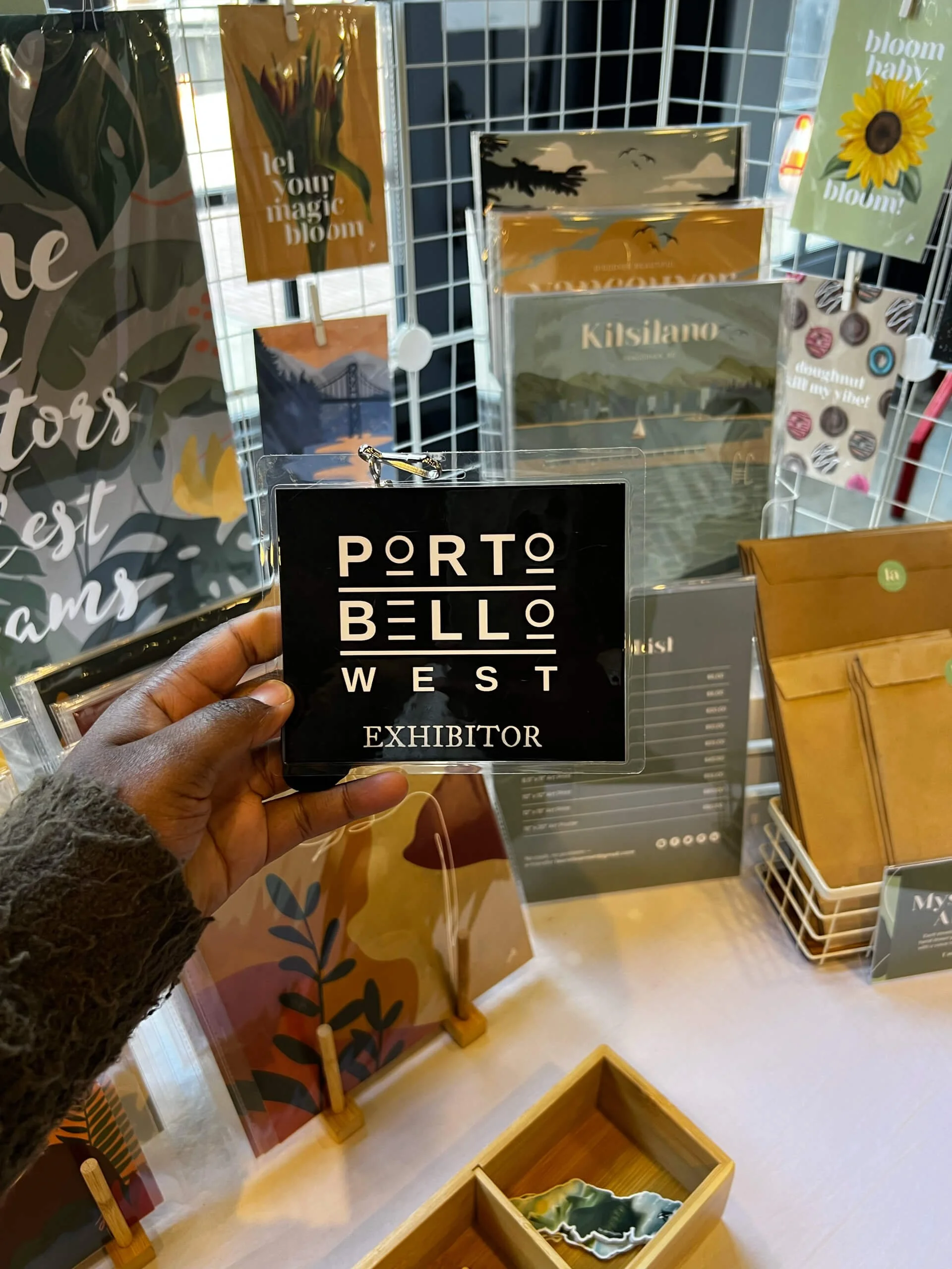 Hand holding a black exhibitor badge with white text that reads "PORTO BELLO WEST EXHIBITOR" against a display of art prints and decorative items at a store or exhibit.