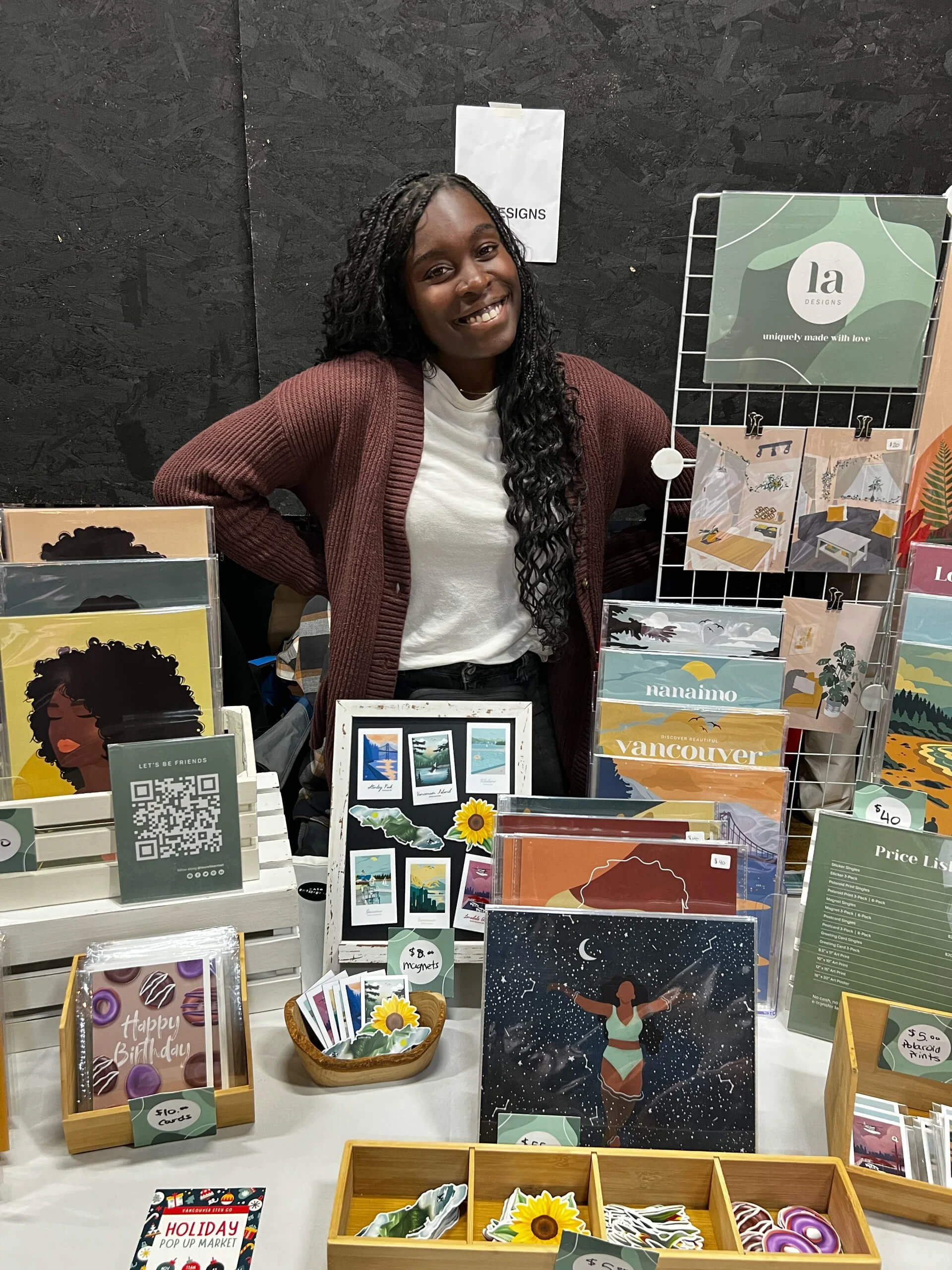 A woman with long curly dark hair and a white shirt wears a maroon cardigan and poses behind a display table of colorful greeting cards and artwork at a craft fair or market. The background is black and textured.