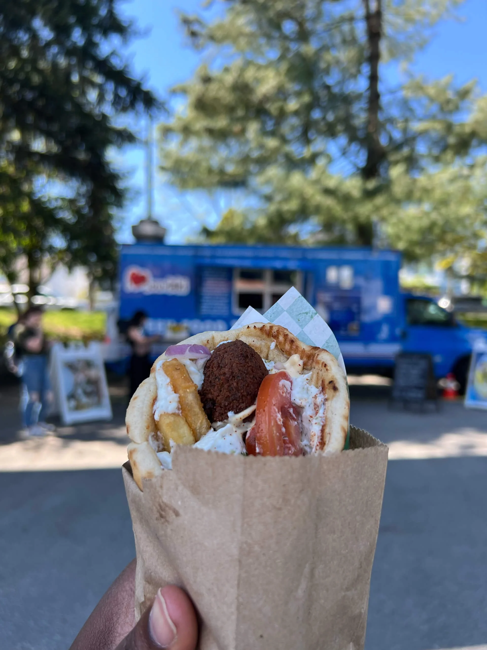 A person holding a slice of pizza with toppings including a meatball, tomato, onions, and cheese, with an outdoor setting in the background featuring trees and a blue food truck.