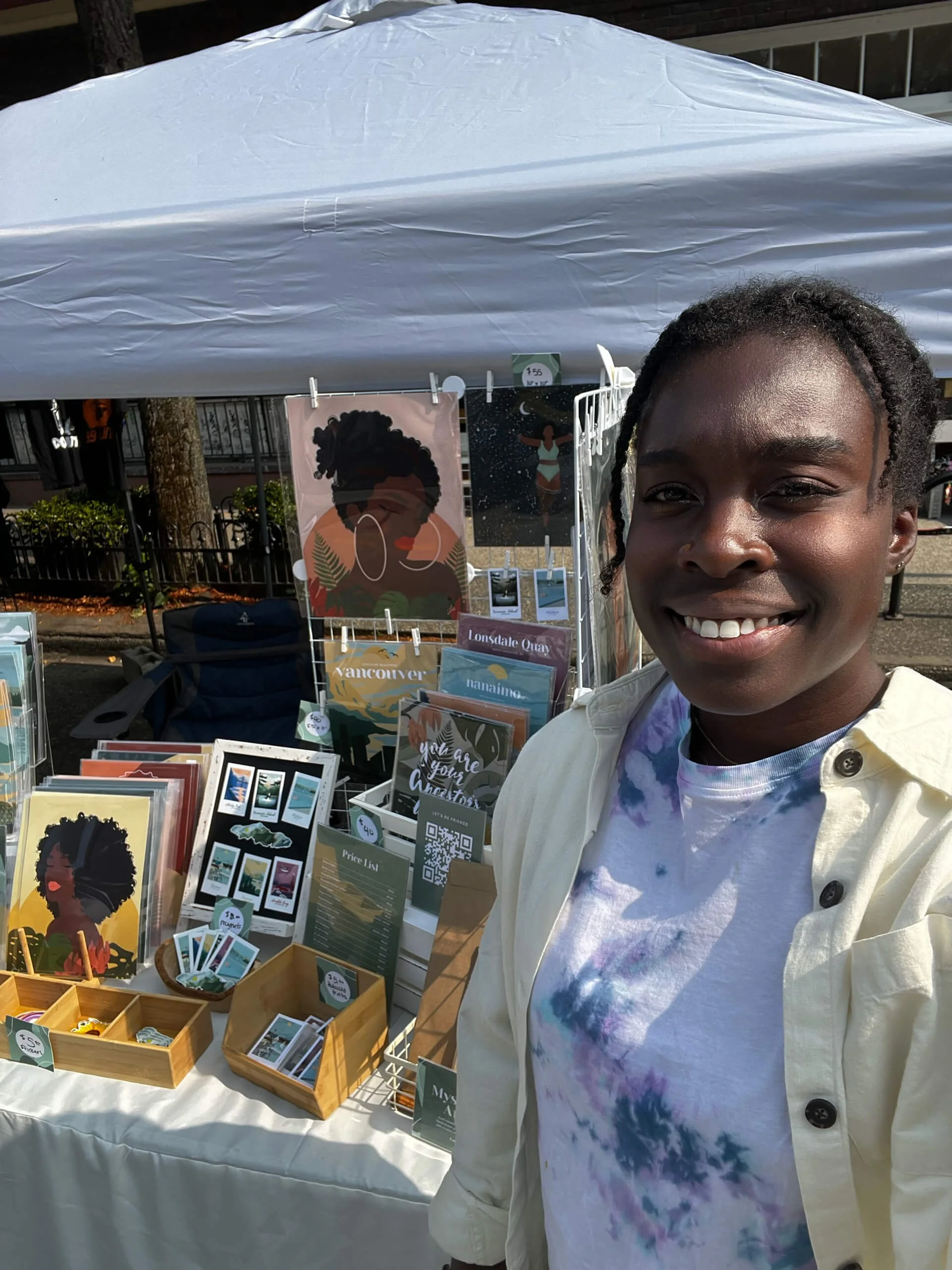 A young woman smiling at an outdoor market booth selling postcards and art prints with illustrations of women and nature scenes. The booth has a white tablecloth and a tent canopy overhead.