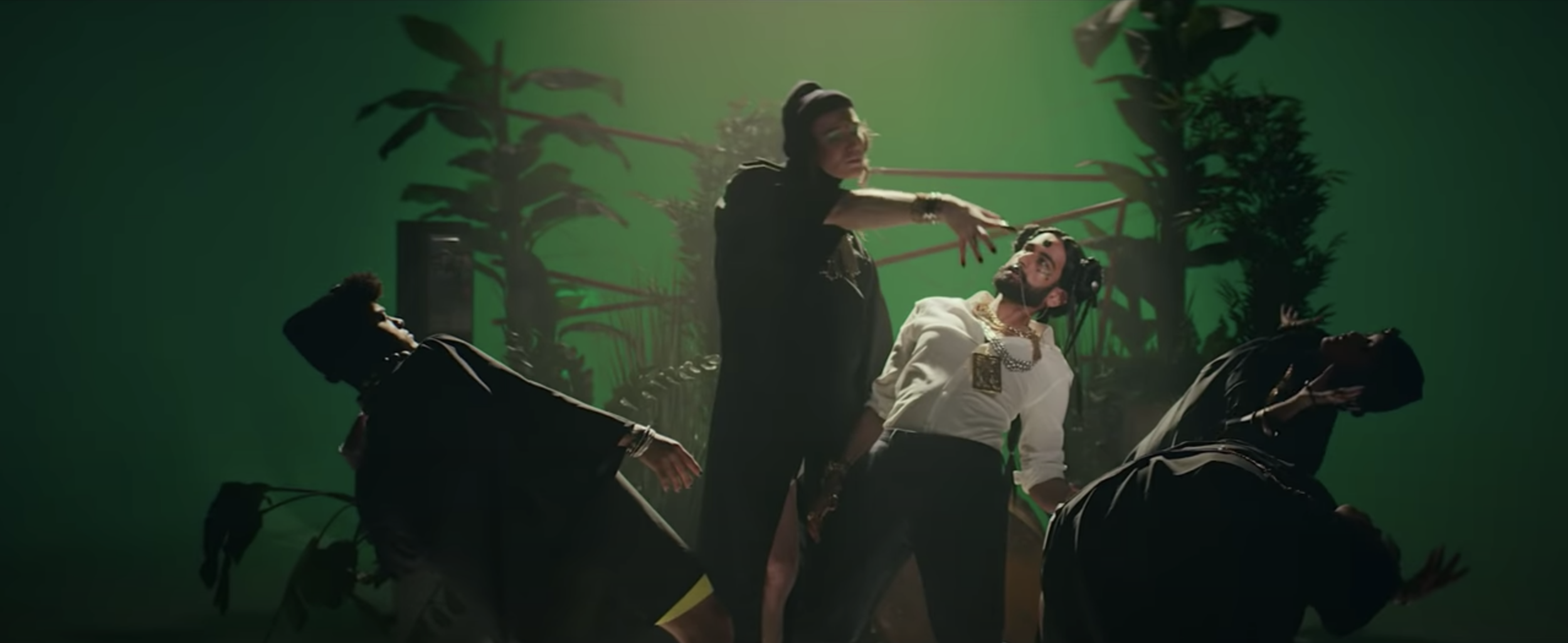 A woman dressed in black with a ponytail and glasses is standing over three men wearing headsets and white shirts, one with a large necklace, in a scene with green screen background and tropical trees.