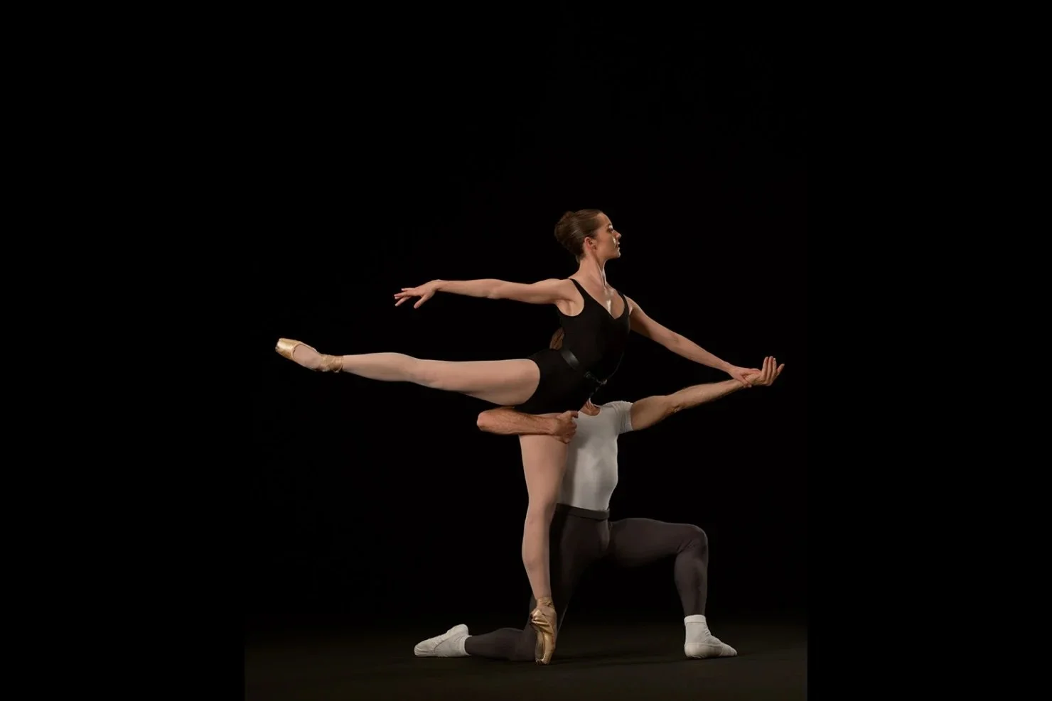 Ballet dancers performing a pose on stage, with one dancer in a black leotard and the other in a grey top and black tights, against a black background.