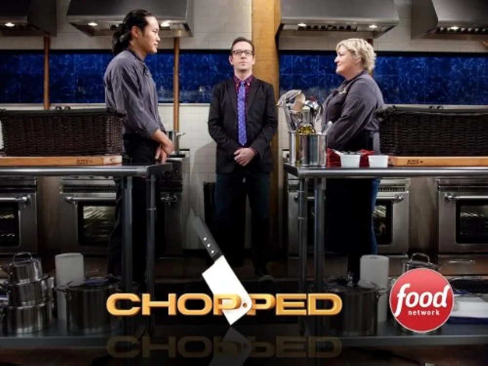 Three people standing in a professional kitchen, two women on the sides and a man in the middle, engaged in a conversation, with the food network logo at the bottom.
