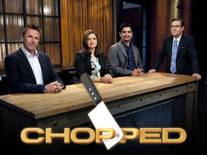 Four TV hosts or judges seated at a wooden table on the set of a show called 'Chopped,' with a large kitchen knife slicing through the show's logo at the bottom.