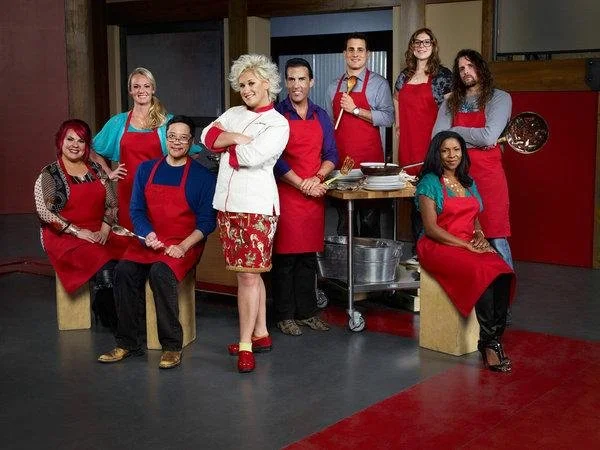 Group of nine diverse people in a kitchen, some sitting and some standing, wearing red aprons and smiling.