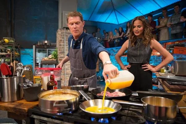 Two chefs cooking on television cooking show, with a male chef pouring eggs into a pan and a female chef observing, with an audience watching in the background.