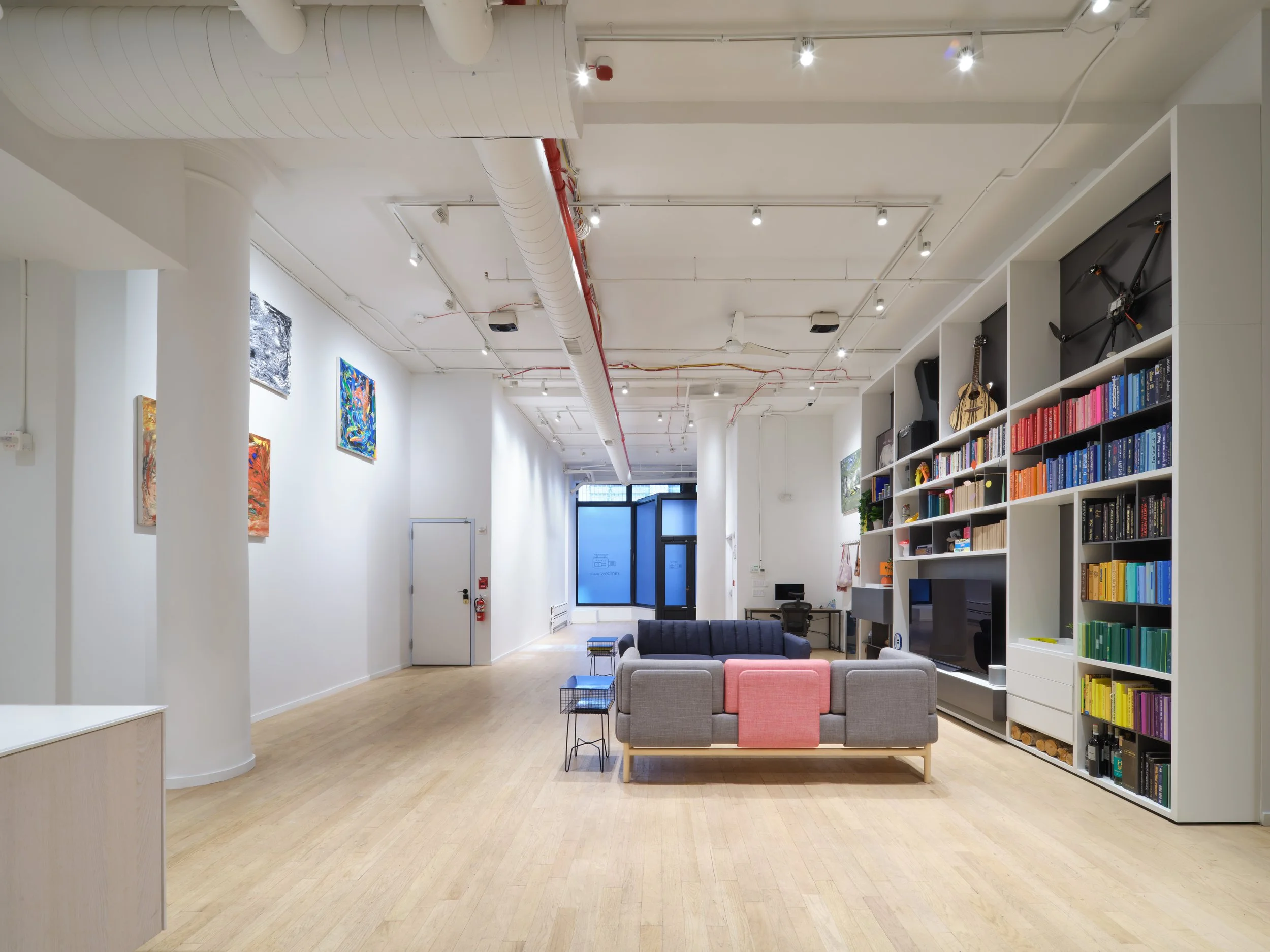 Modern interior of an art gallery or studio with white walls, hardwood floors, and colorful paintings on the left wall; open space with seating area including two sofas and a small table; a large white shelving unit on the right filled with books and