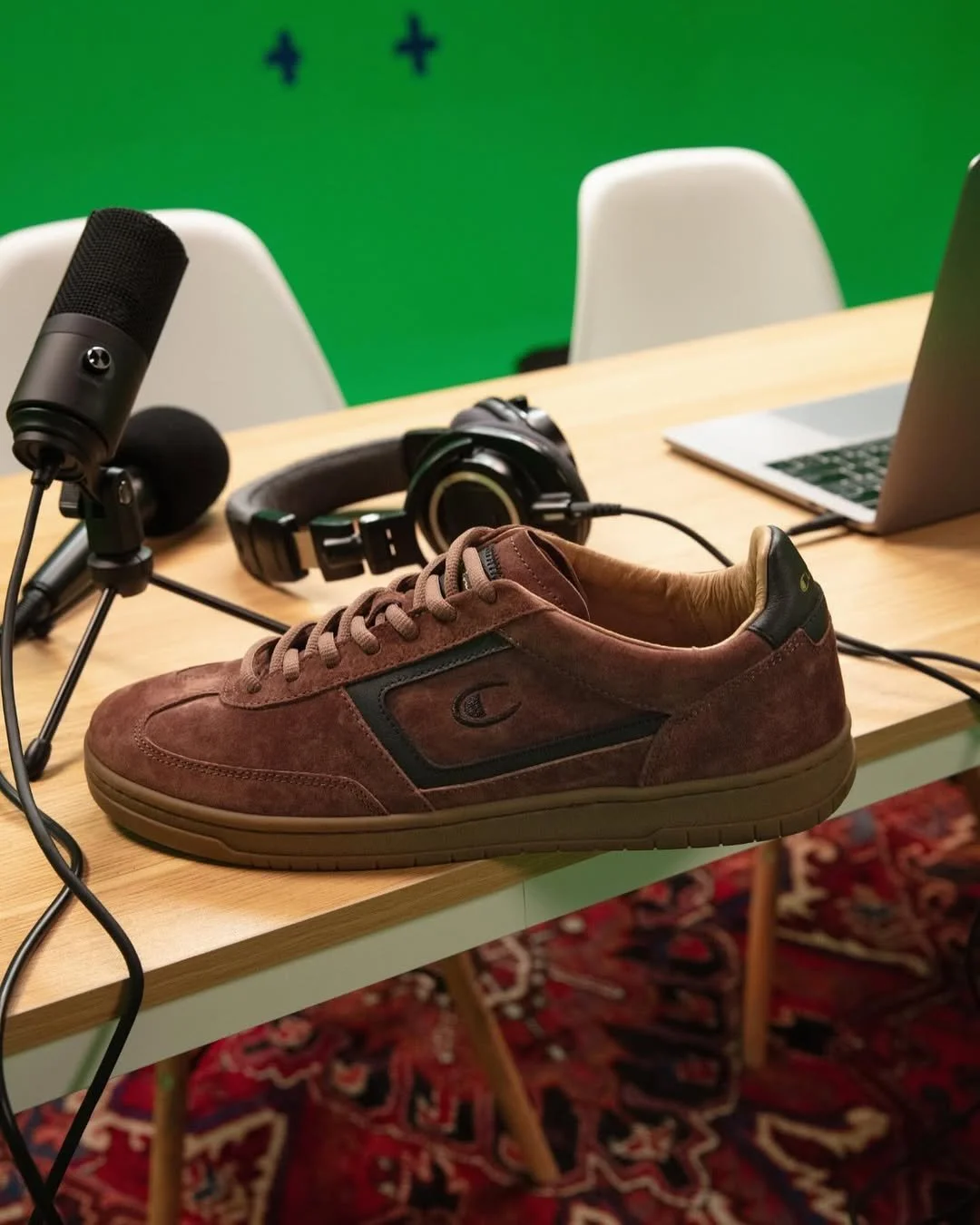 A brown athletic shoe with black accents on a wooden table in a podcast recording studio with a microphone, headphones, a laptop, and a green backdrop.