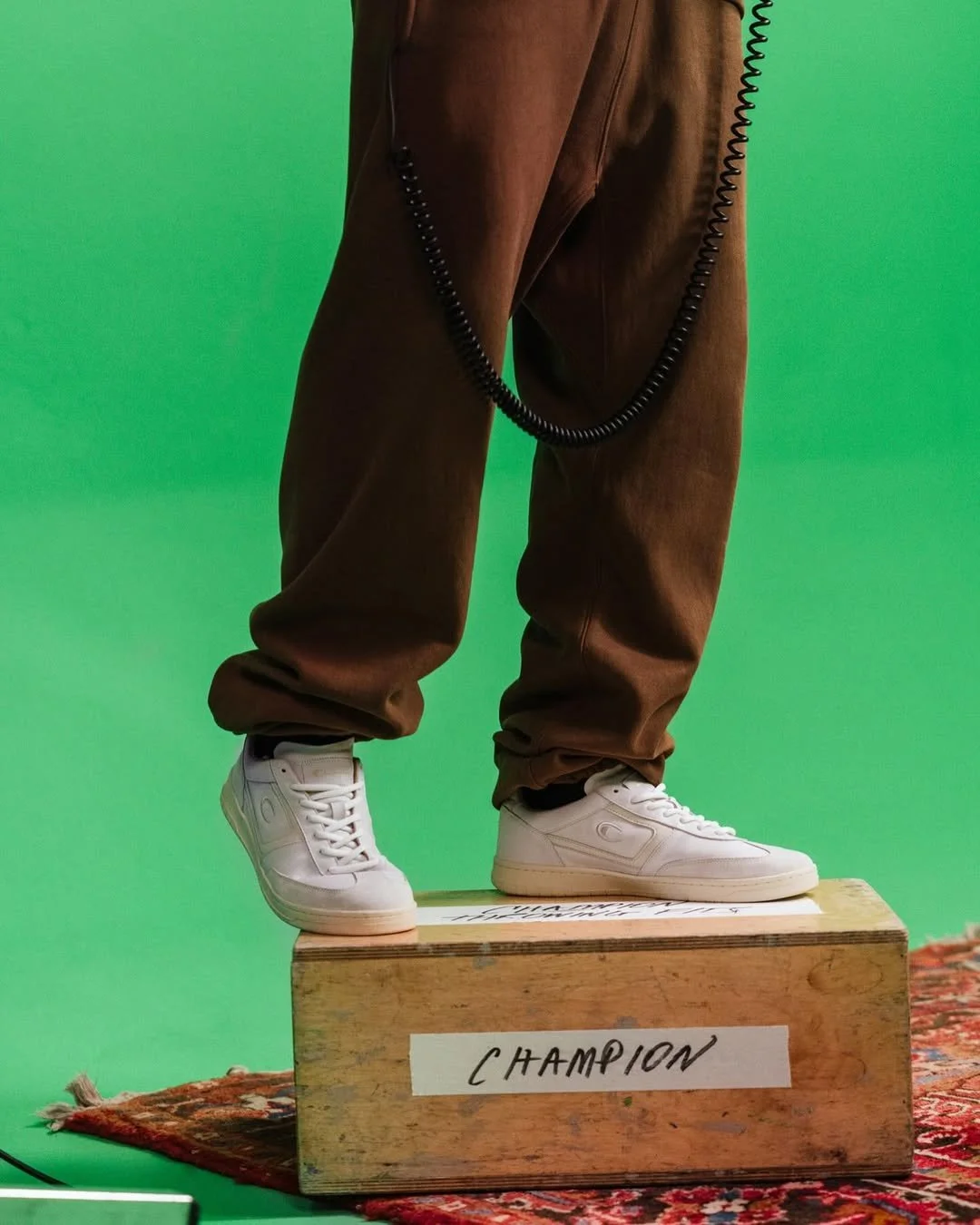 Close-up of a person standing on a wooden box labeled 'Champion' against a green background, wearing brown pants and white sneakers, with a black coiled wire hanging from their waist.