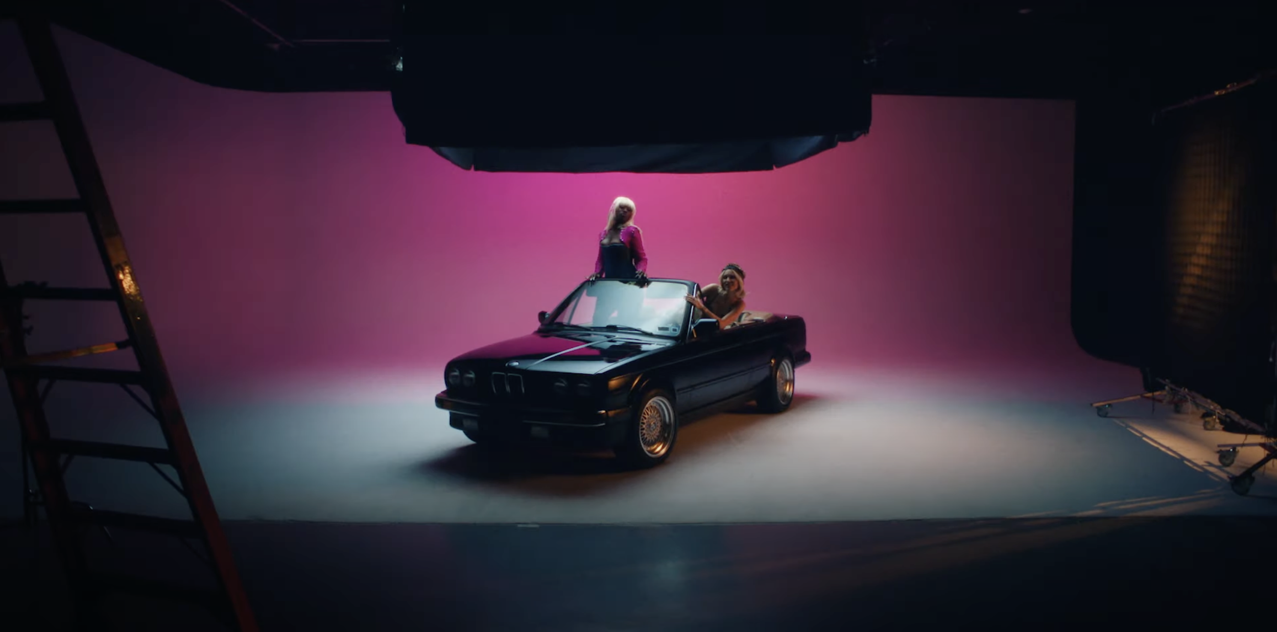 Two women in a black convertible car in a photography studio with pink backdrop, studio lights, and equipment visible.