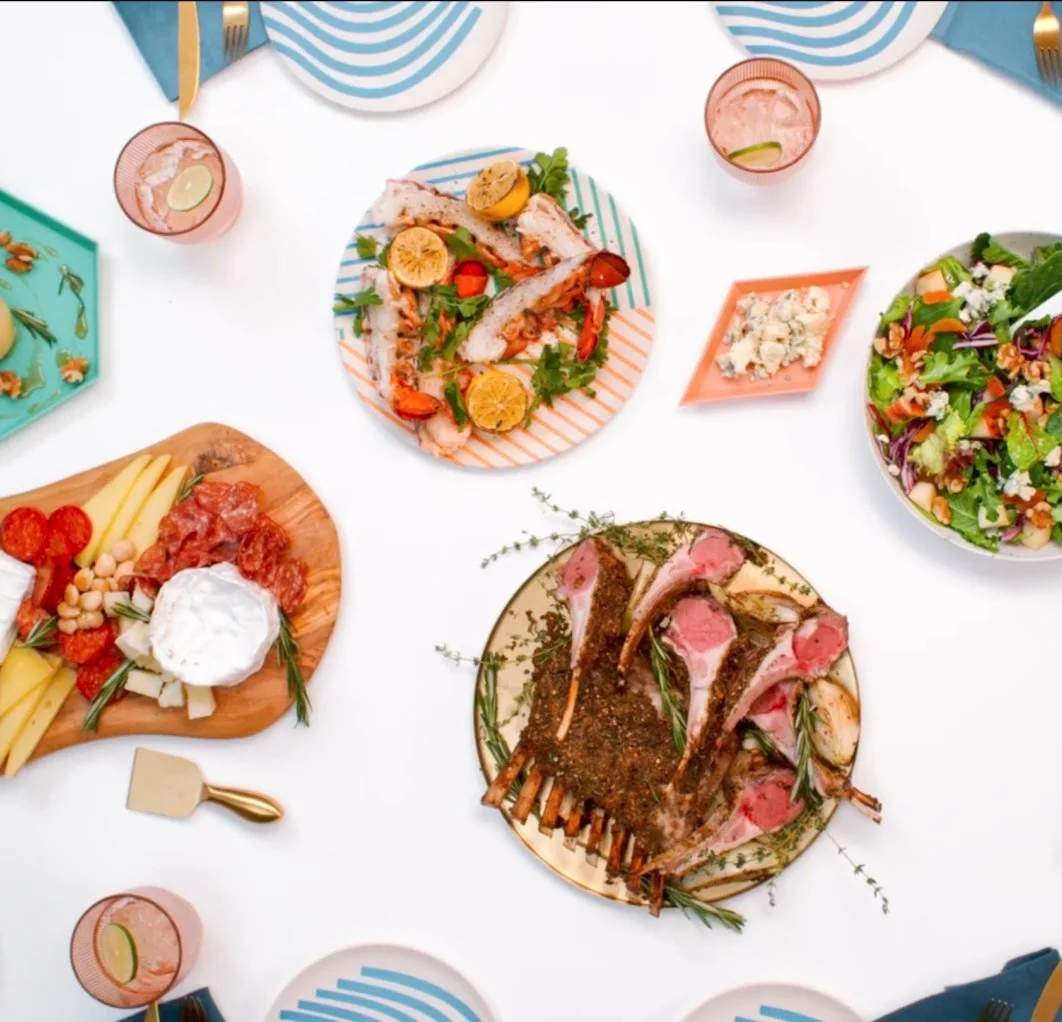 A table set for a meal with various dishes including a green salad, grilled lamb chops, cheese and charcuterie platter, a fish dish with lemon slices, and drinks in pink glasses with lime slices.
