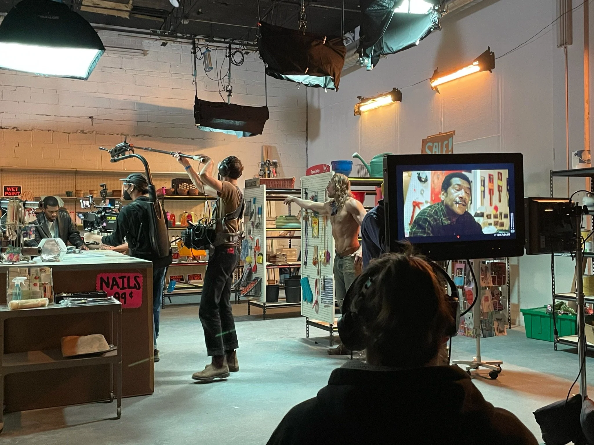 Behind the scenes of a film or TV production in a workshop or store setting, with crew members preparing equipment. A shirtless actor with long hair points or gestures as a person dressed as a character with a fake mouth and teeth looks on. A monitor