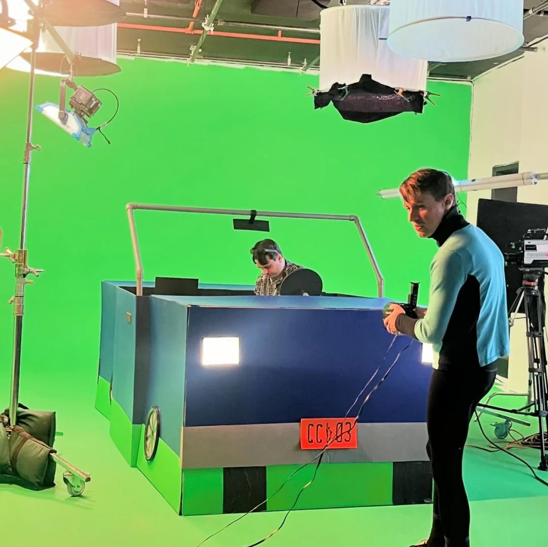 Film studio with a large green screen backdrop, lighting equipment, and two crew members preparing for filming; one person is operating a camera, and another is working on a monitor.