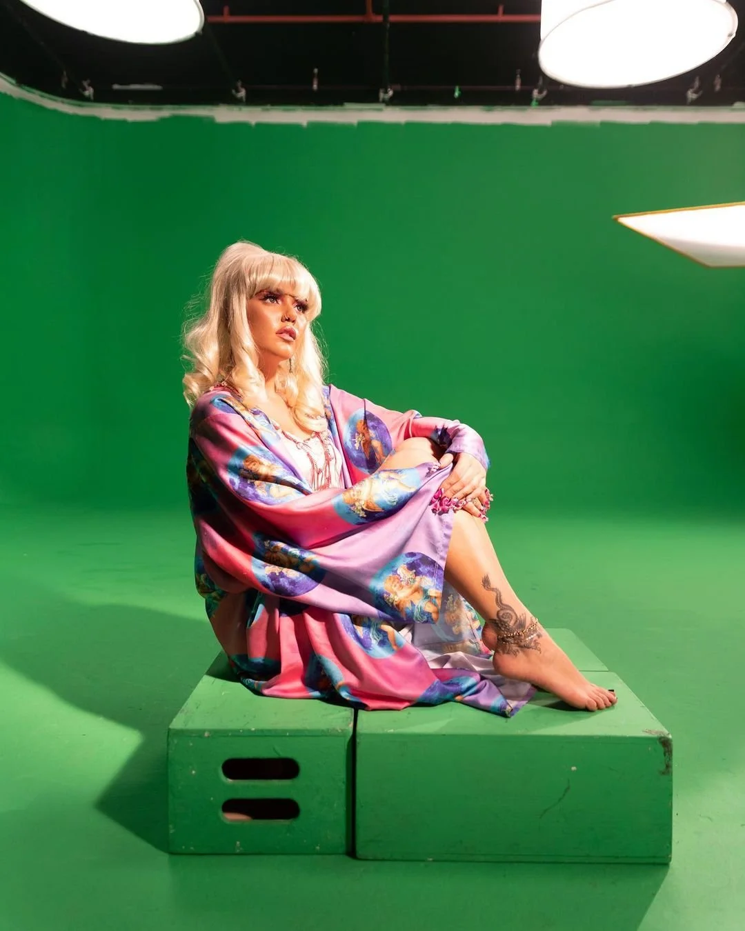 A woman with blonde hair sits on a green platform in front of a green screen, wearing a colorful silk robe and looking contemplative.