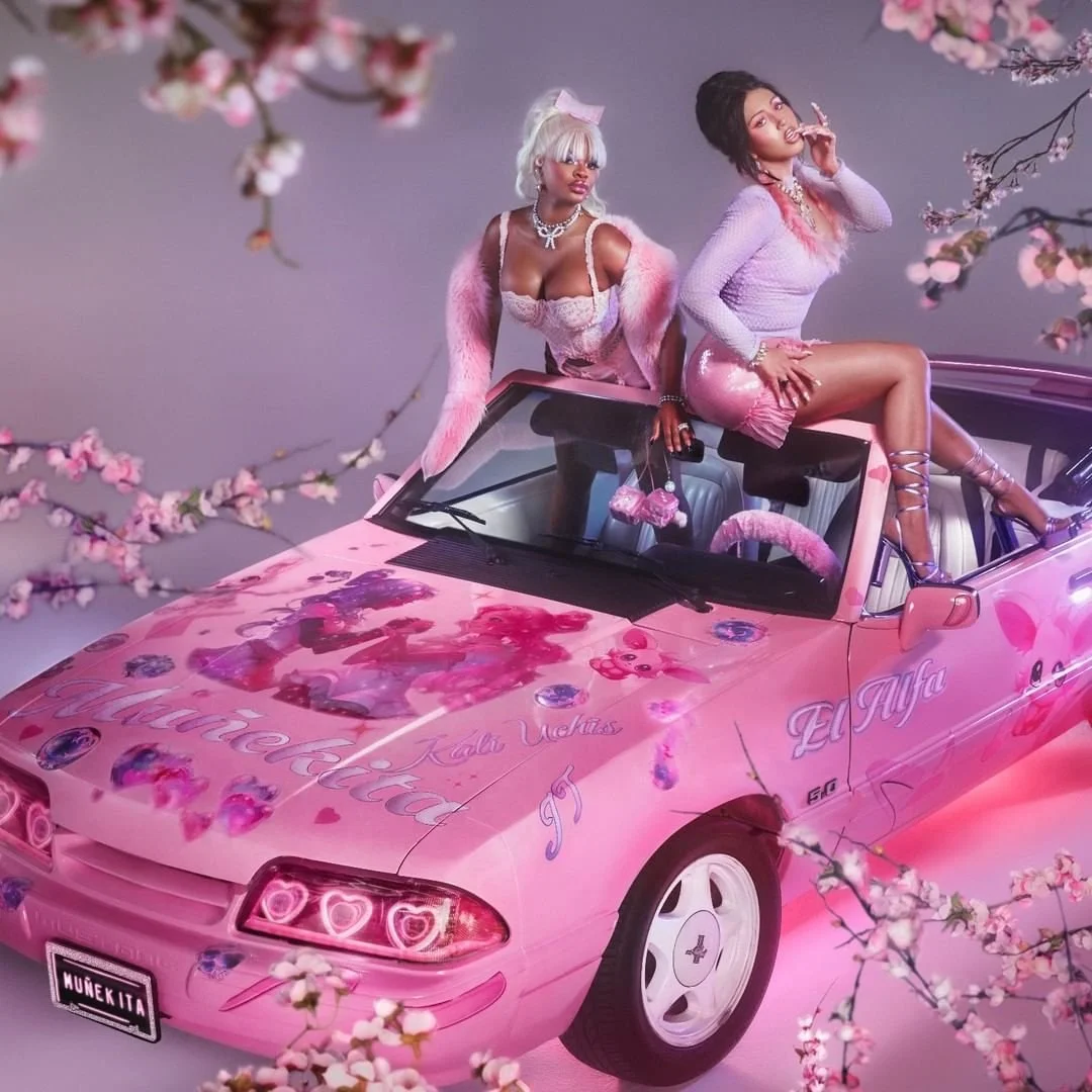 Two women dressed in pink and purple clothing, with one sitting on the hood and the other sitting on the windshield of a pink convertible car decorated with heart-shaped lights and floral patterns, surrounded by pink flowering branches.