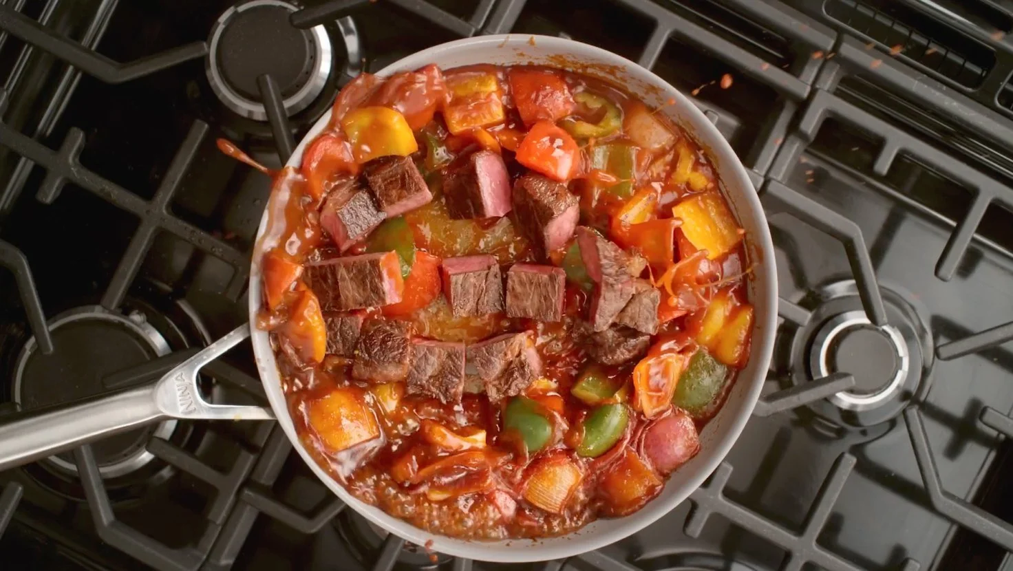 A skillet on a stove filled with stew containing chunks of beef, bell peppers, tomatoes, and onions in a tomato-based sauce.