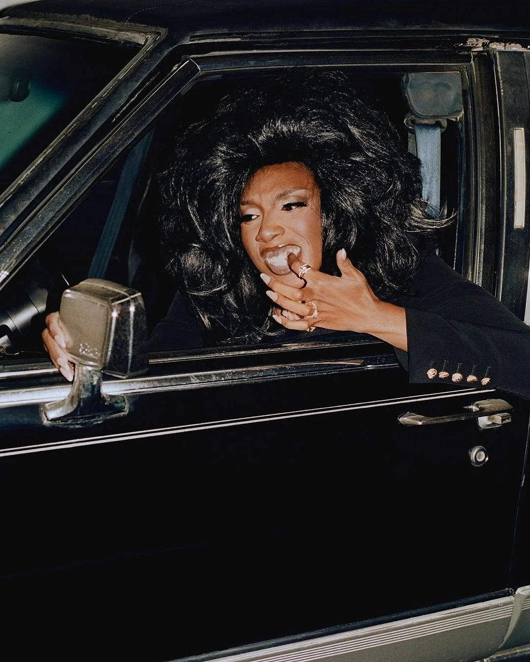 A woman with voluminous black hair sitting in a vintage black car, looking distressed and biting her fingers.