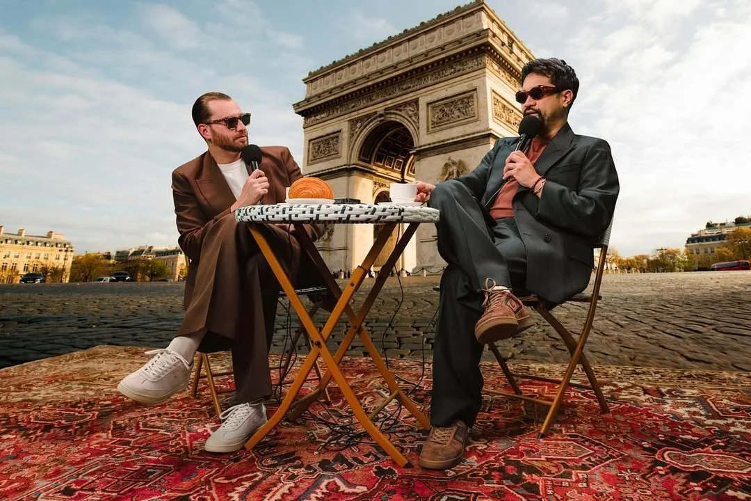 Two men with sunglasses sit at a small table with a patterned tablecloth and a croissant, in front of the Arc de Triomphe in Paris. They are holding microphones and appear to be having a conversation.