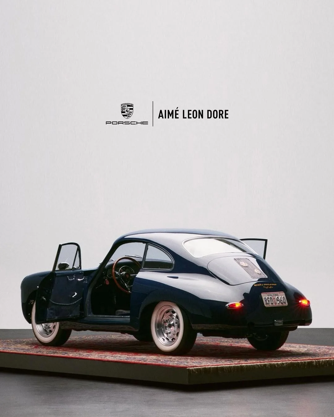 A miniature model of a vintage Porsche car with ultra-wide white tires, dark blue body, open driver's side door, and wooden steering wheel, placed on a small rug against a white background featuring Porsche logo and the words 'Aimé Leon Dore'.