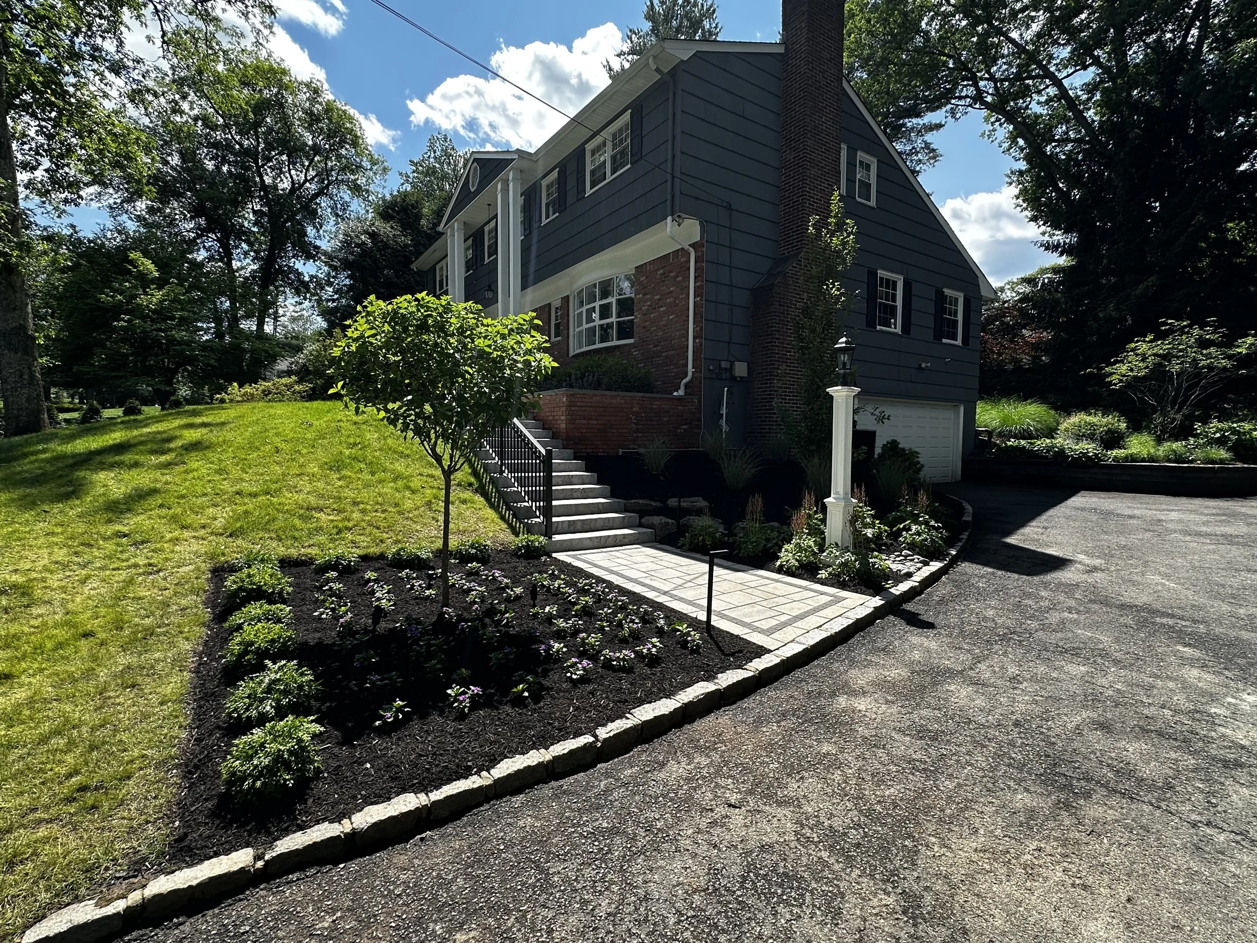 Front yard of a blue house with porch and stairs, landscaped garden with small tree and flowers, paved driveway, tall trees, partly cloudy sky.