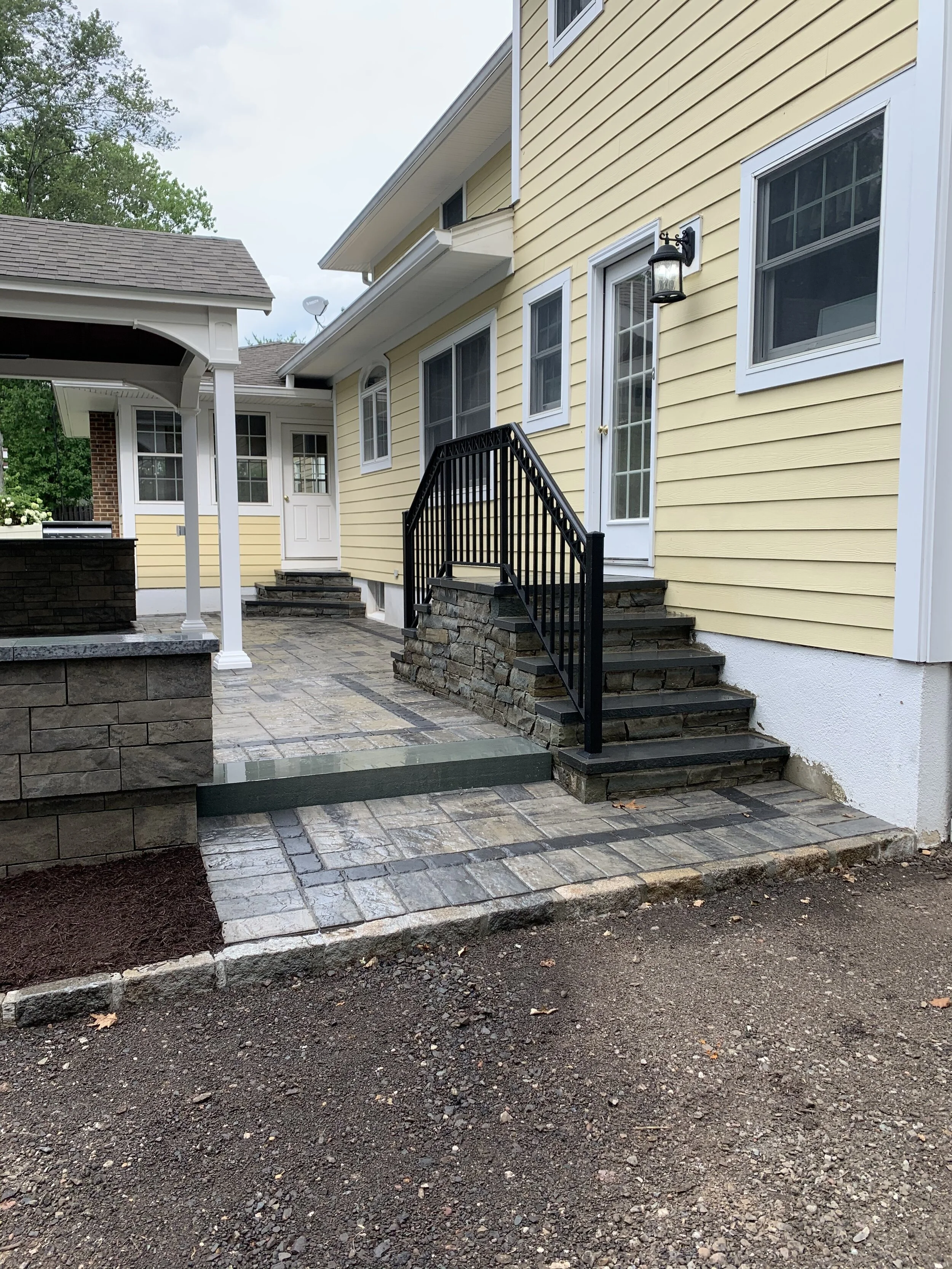 A newly constructed backyard patio with stone pavers, black metal railing stairs, yellow siding house with multiple windows and a glass door, and an outdoor light fixture.