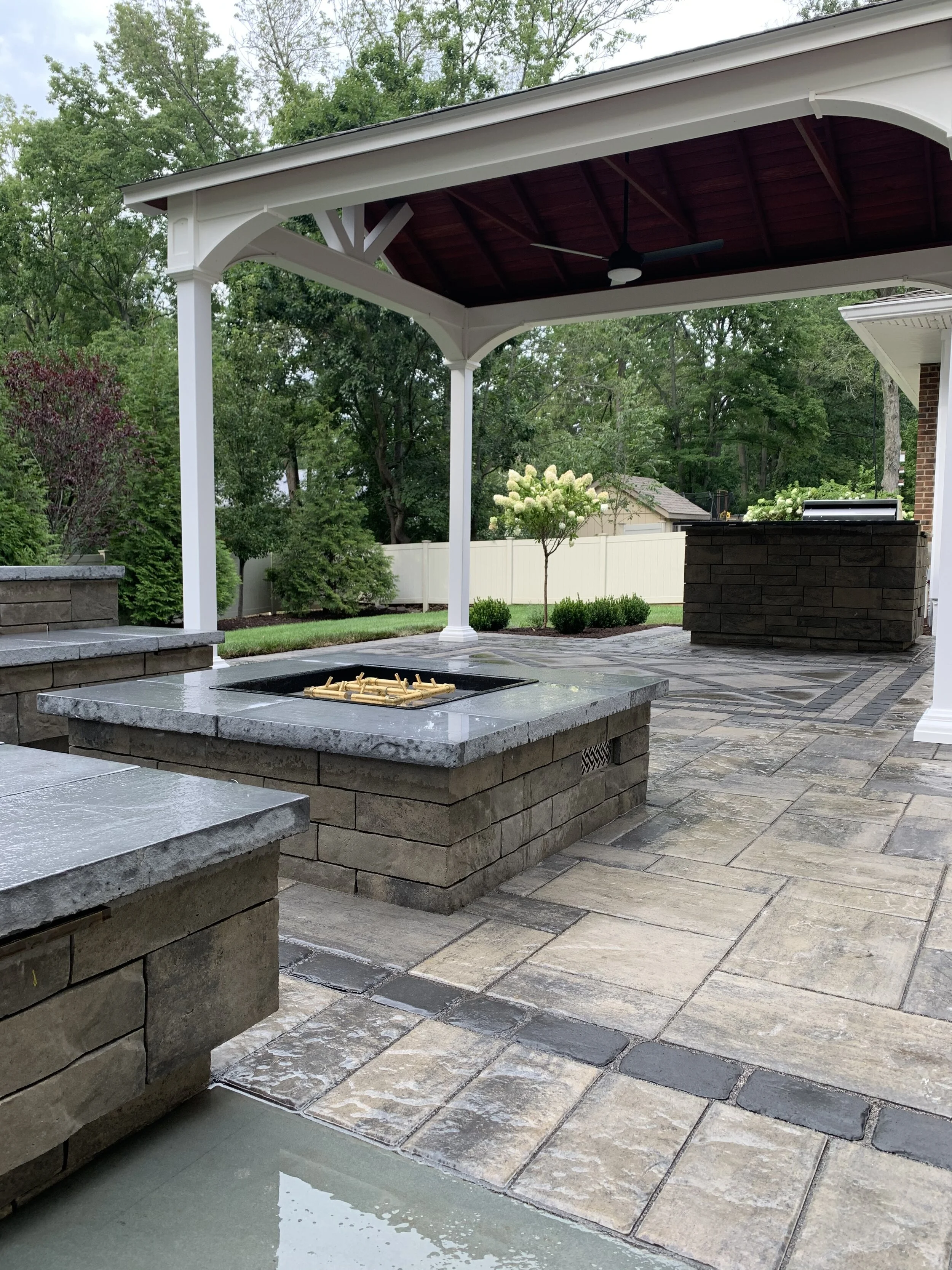 Backyard patio with stone flooring, built-in firepit, covered seating area, ceiling fan, and lush green trees in the background.