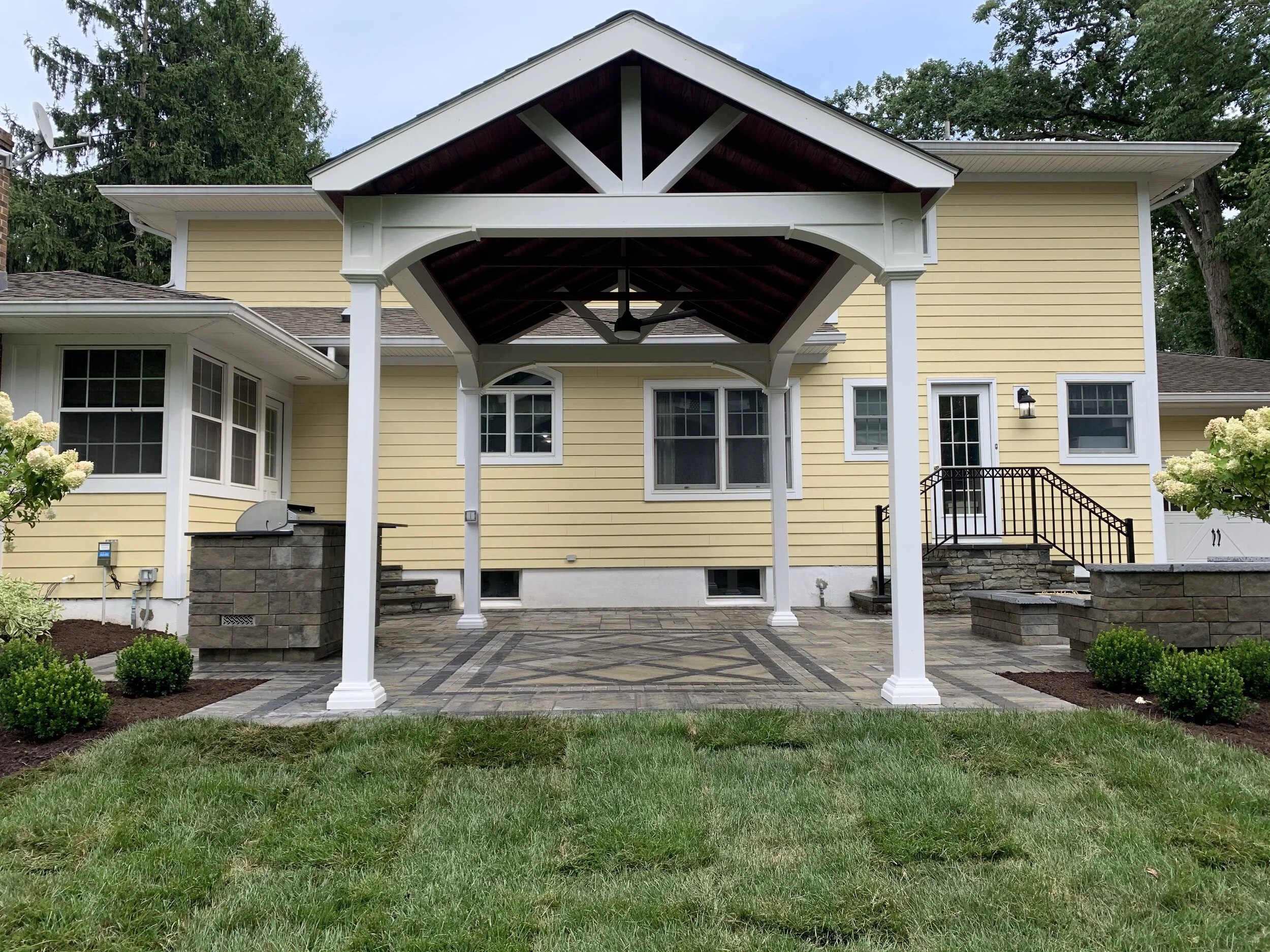 Backyard patio with a covered wooden pergola, stone paver flooring, and yellow house with multiple windows and stairs leading to the door, surrounded by landscaped plants and grass.