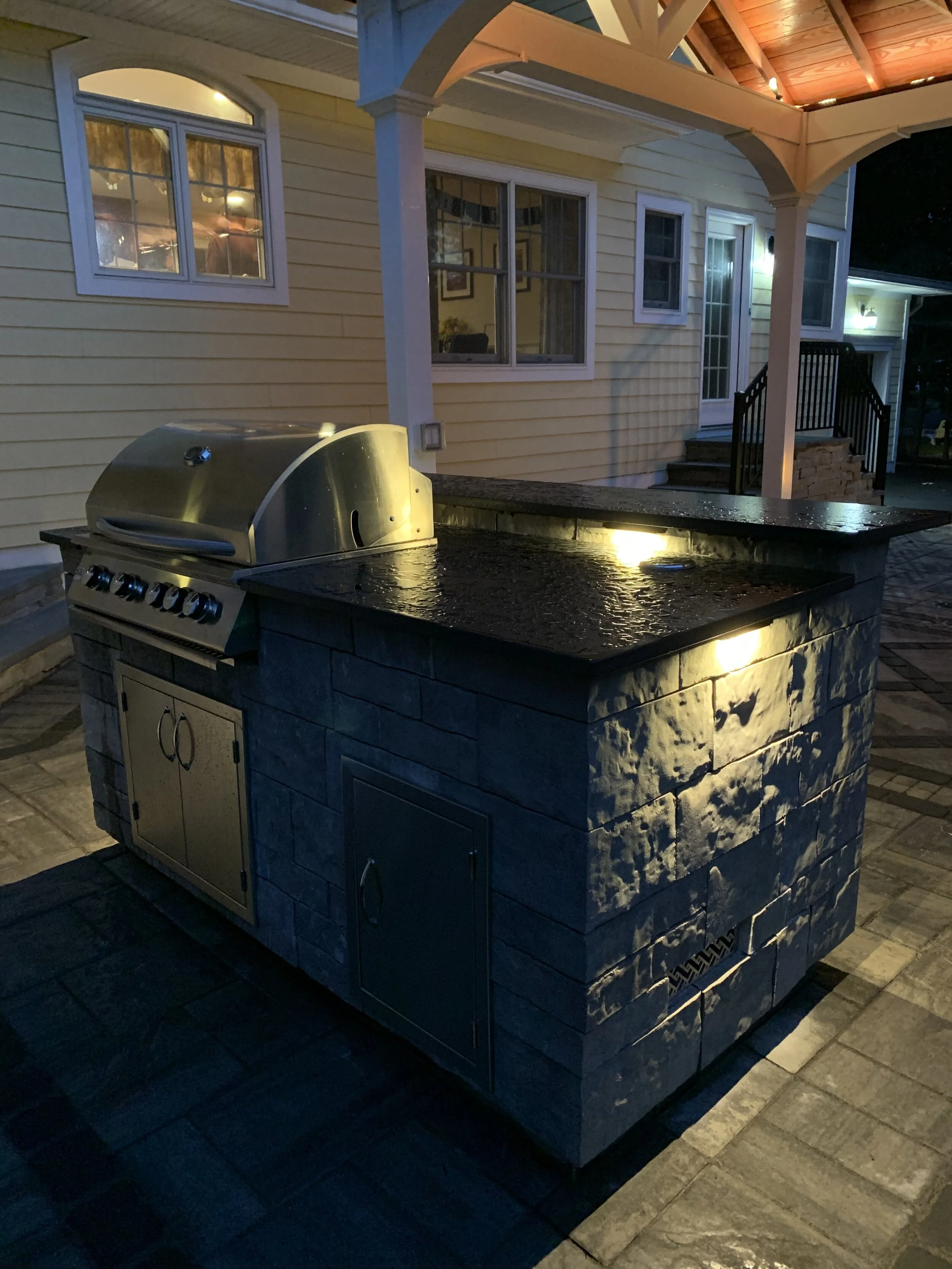 Outdoor kitchen with a built-in grill on a stone island at night, illuminated by lights under the roof of a patio.