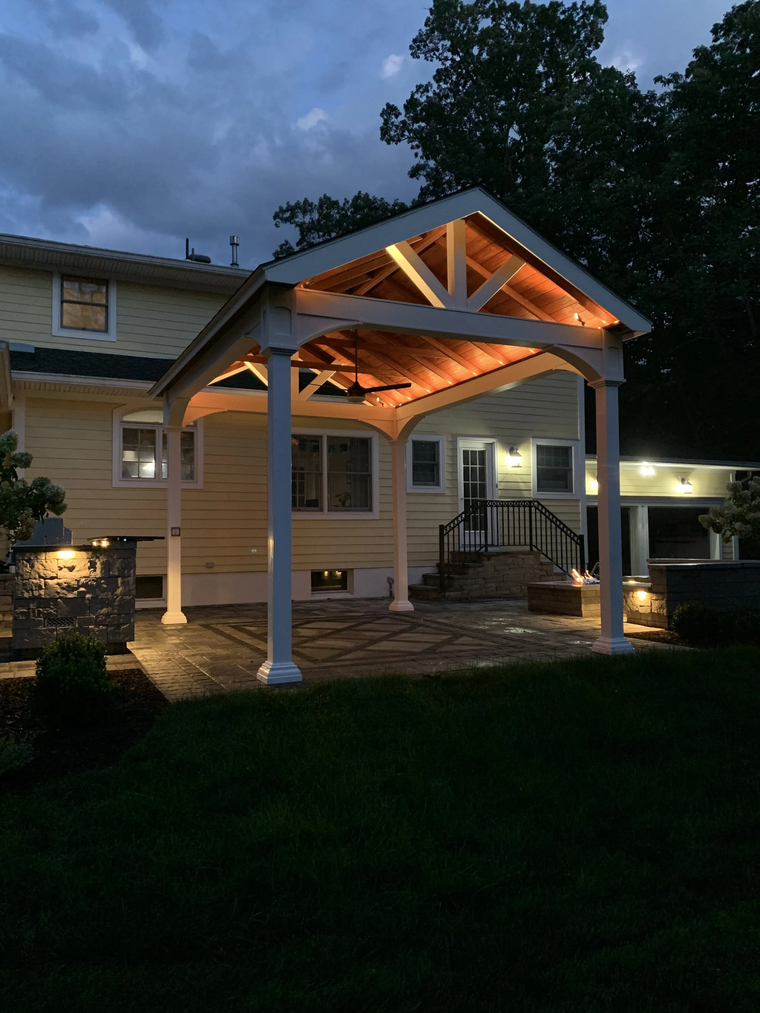 Backyard patio with a covered pergola, string lights, and outdoor fireplace at dusk.