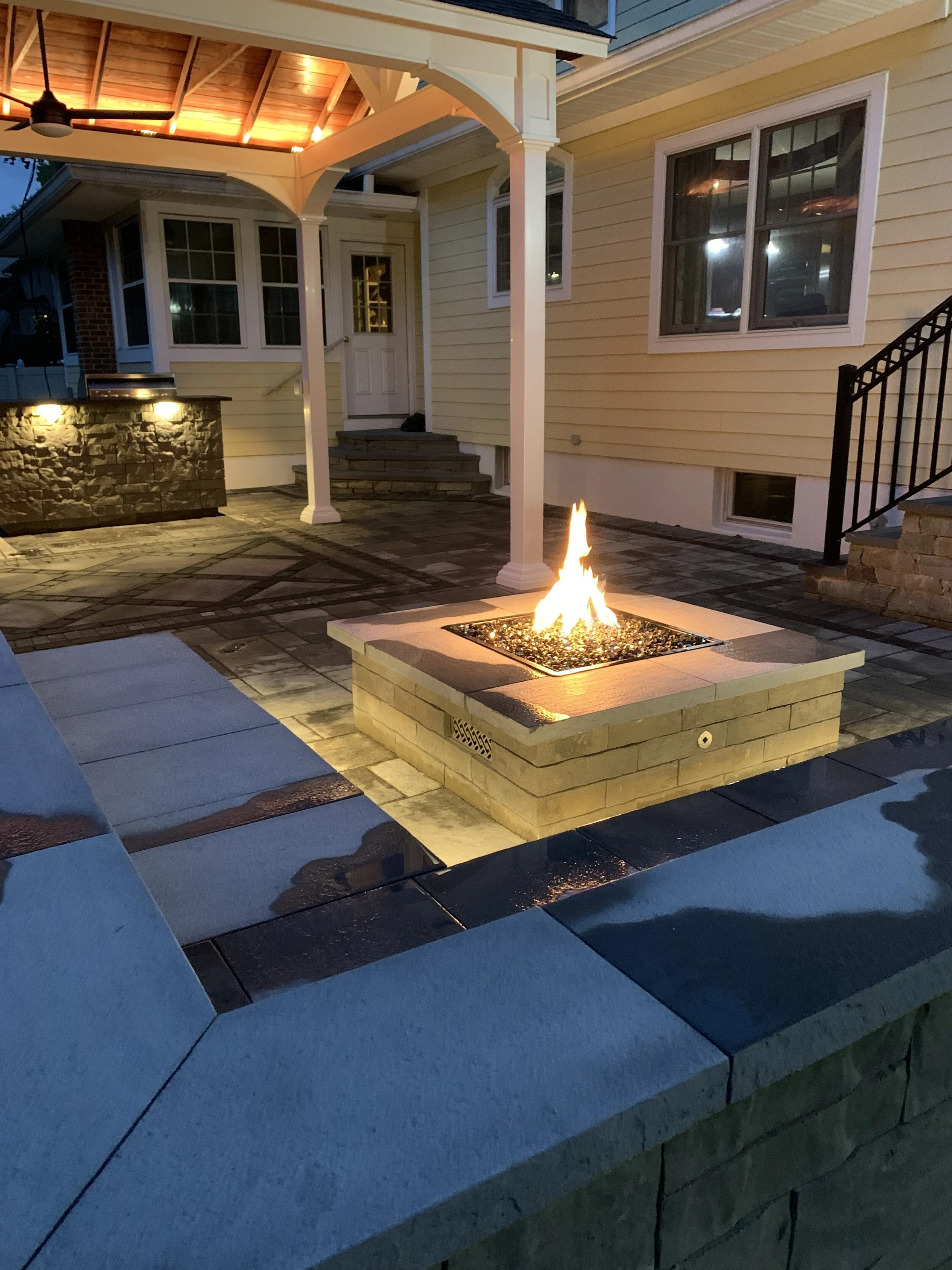 Outdoor patio with a fire pit in the center, surrounded by seating, and a house with stairs, windows, and a covered porch.