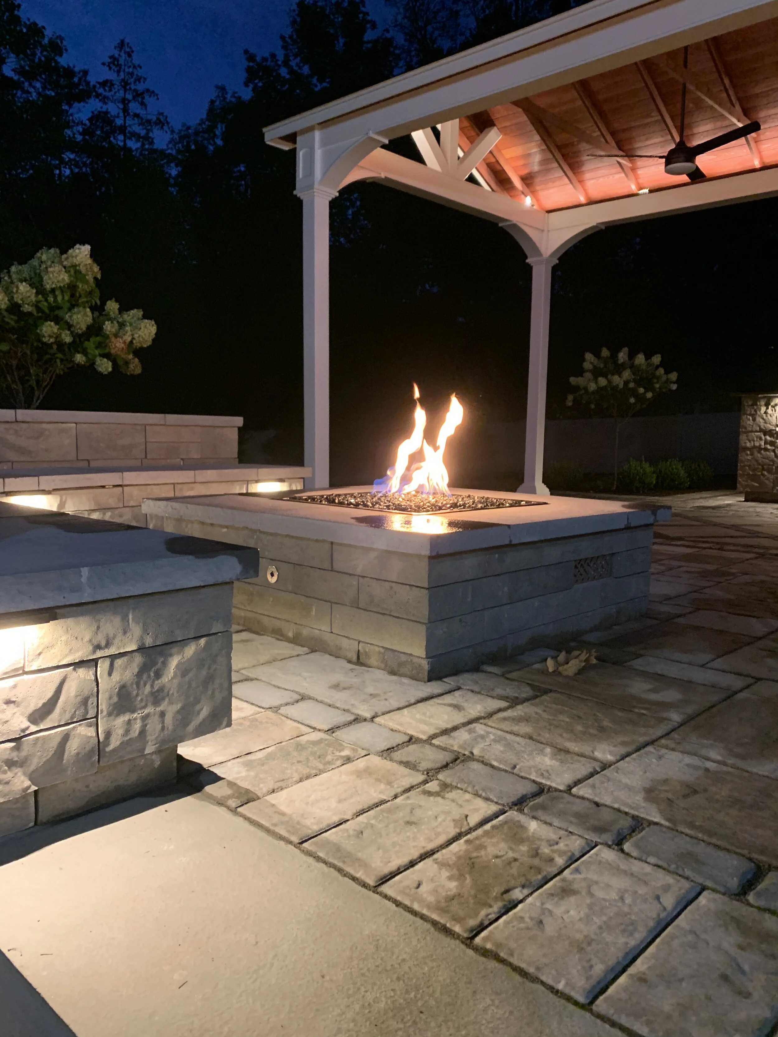 An outdoor fire pit at night within a stone patio area, with a covered pavilion, trees, and flowering bushes in the background.