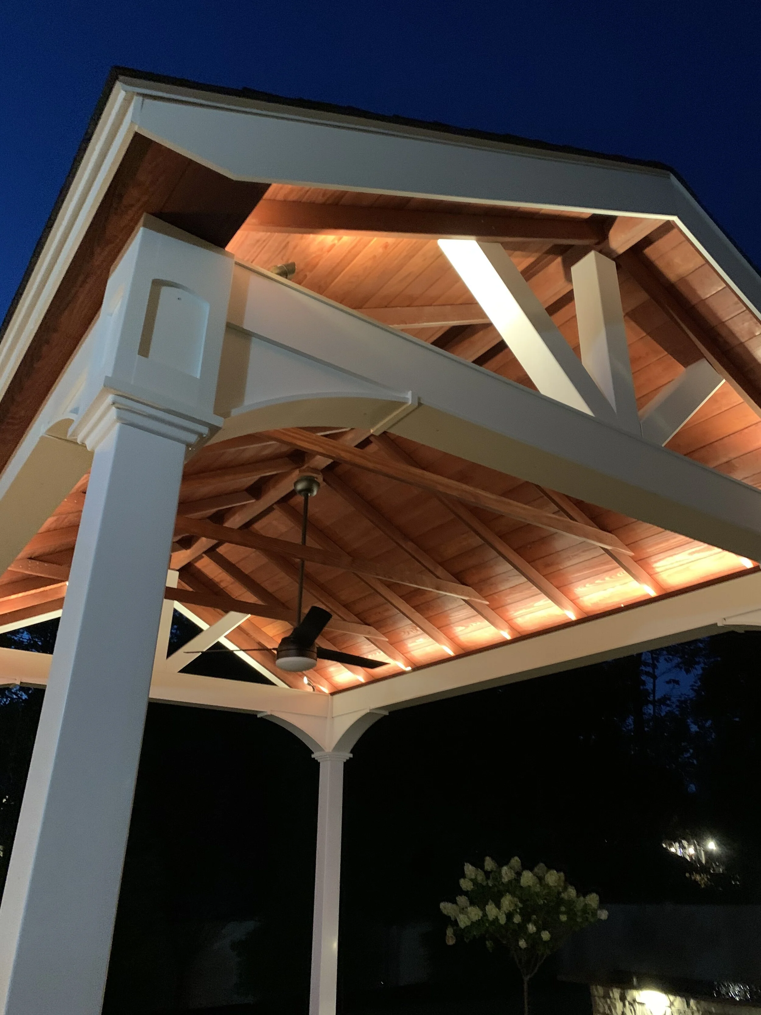 Under a peaked wooden roof supported by white columns, a ceiling fan hangs from the ceiling, which is lit by warm lighting at night.