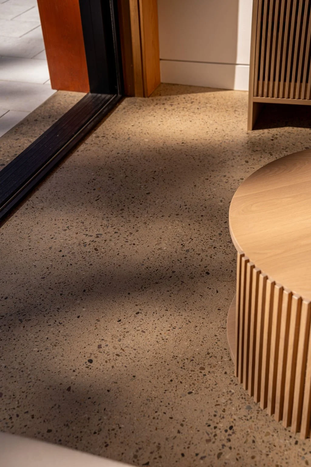 Part of a room with a speckled floor, a round wooden table, a wooden slatted divider, and a door threshold leading to another room with tile flooring.