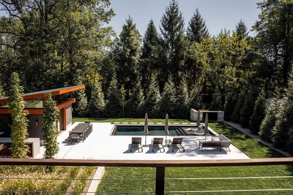 A backyard with a pool, lounge chairs, umbrellas, a fire pit, and surrounding trees and greenery.