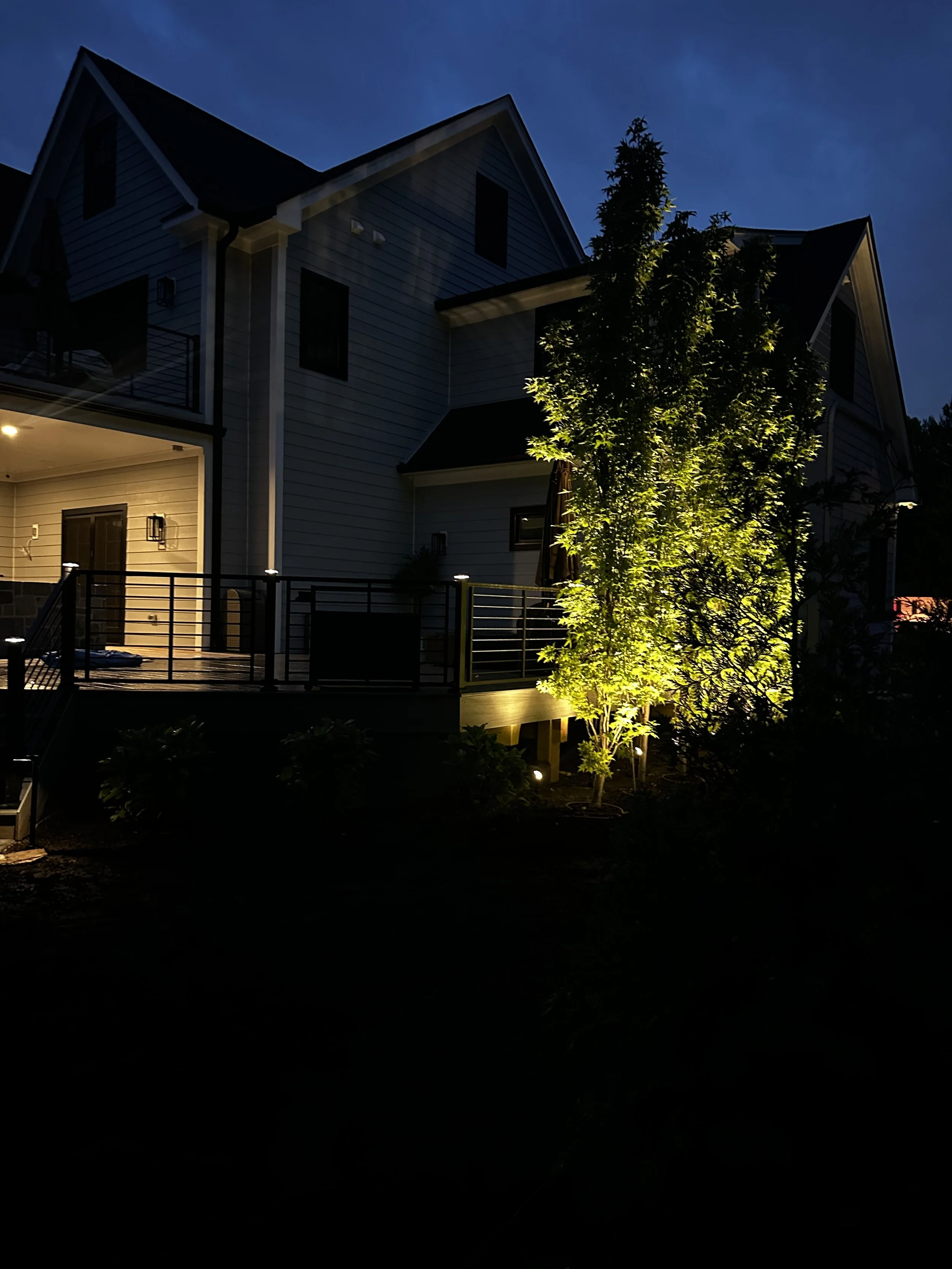 A two-story house at night with exterior lights on, a deck with a railing, and illuminated trees in the yard.