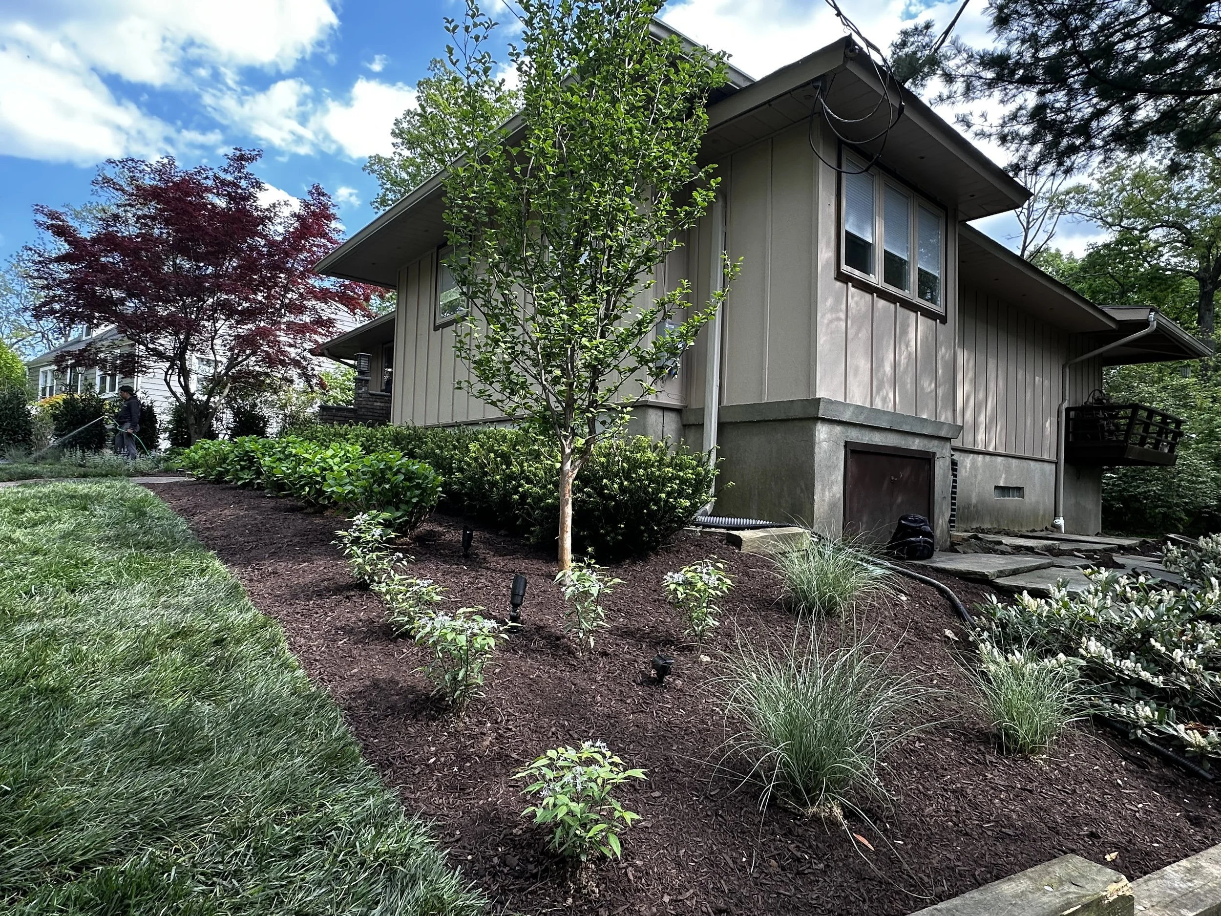 Side view of a house with a landscaped garden, small trees, shrubs, and a person watering plants under a partly cloudy sky.