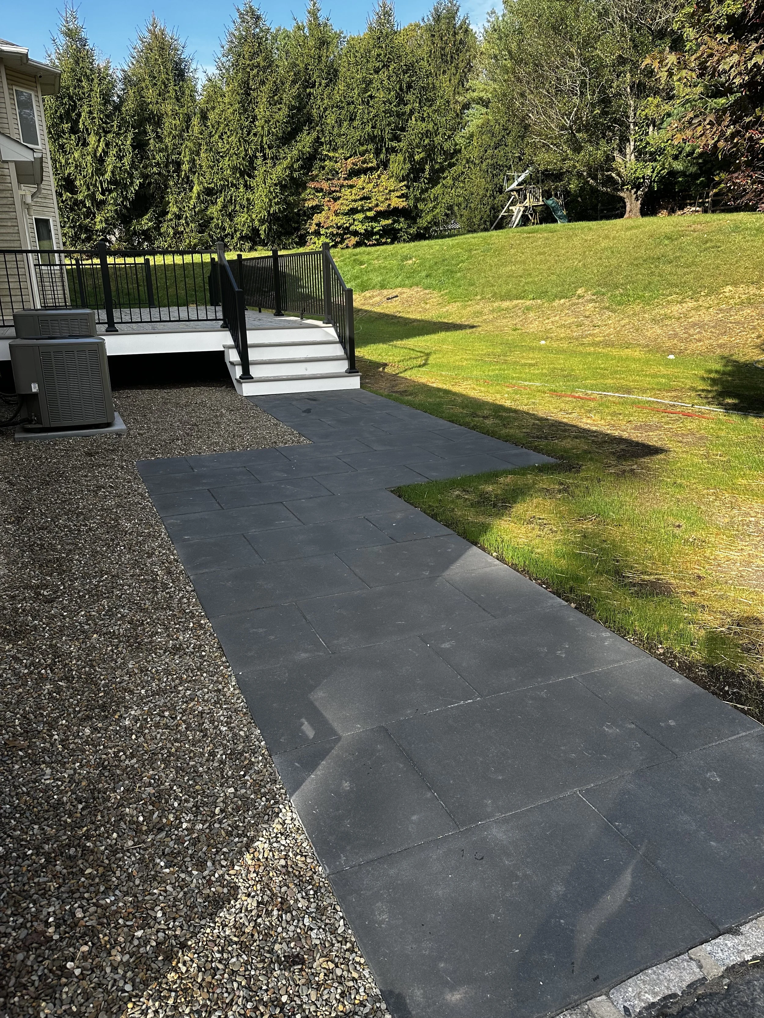 Backyard patio with a paved walkway, a small deck with stairs, and surrounding grassy yard with trees in the background.